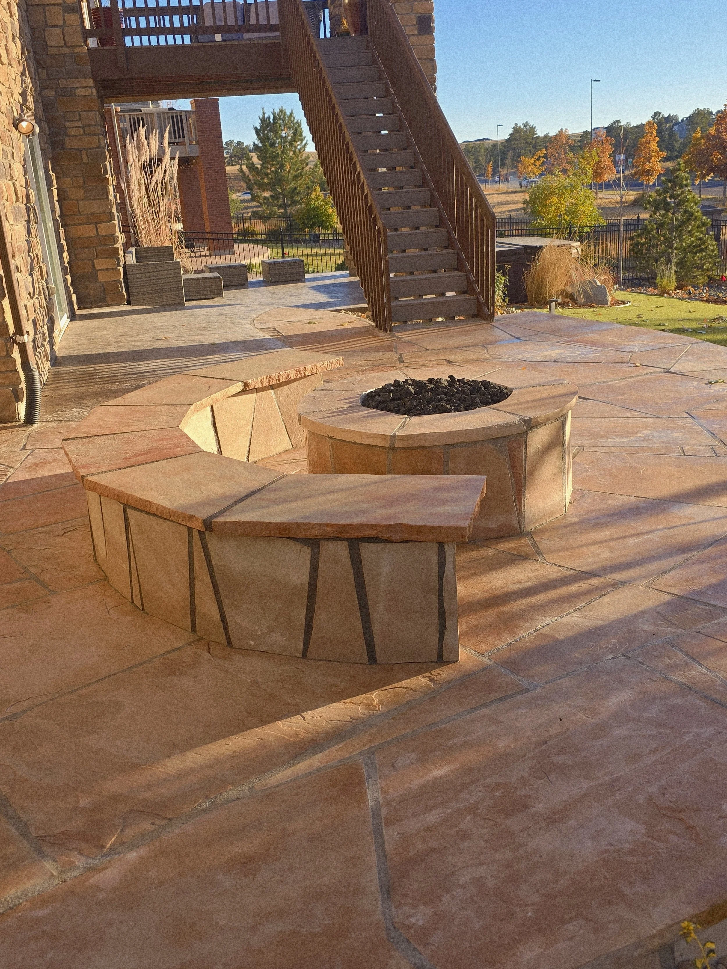 An outdoor patio with a circular fire pit made of brick and a curved stone bench. The patio has large sandstone tiles and there is a wooden staircase leading up to a second level. In the background, there are trees with autumn-colored leaves, a lawn, and a black metal fence.