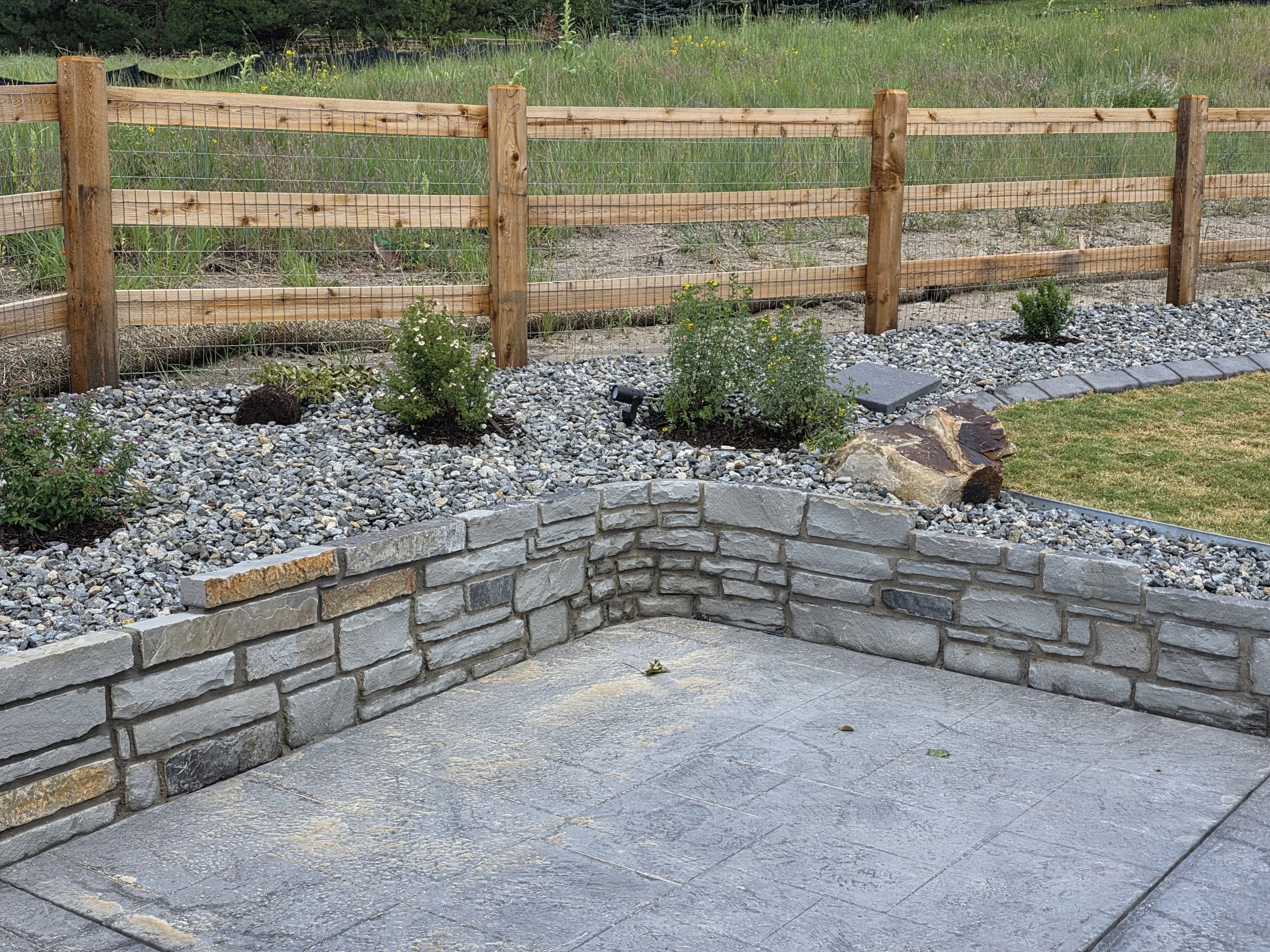 A landscaped outdoor area with a grey stone patio, a short stone wall, and a garden bed with small bushes and gravel, enclosed by a wooden fence. There's a grassy area and a grassy field beyond the fence.