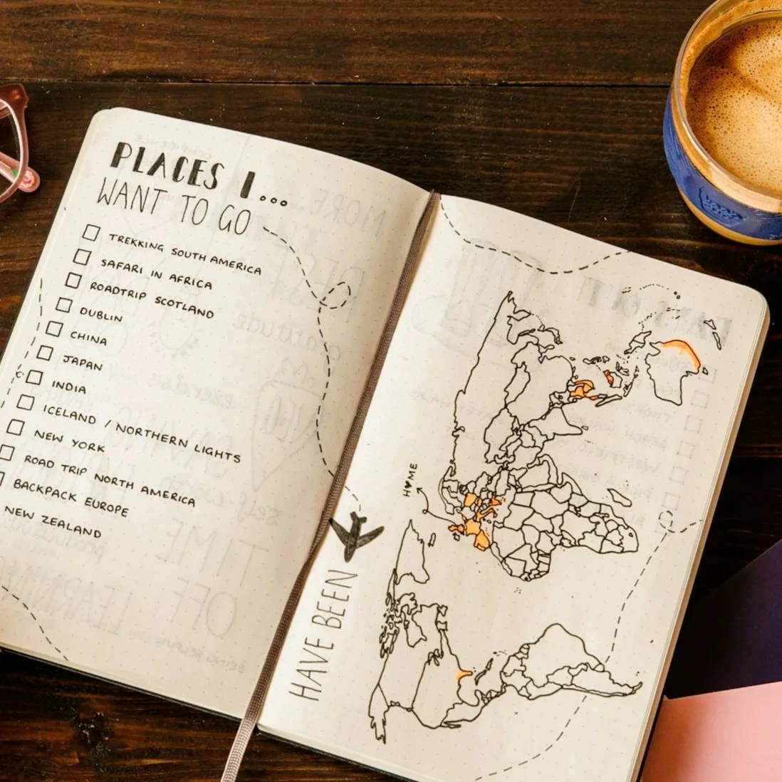Open journal on a wooden table with a travel bucket list and world map. "Places I Want to Go" is listed on the left, including destinations like South America and Africa. The right page shows a map with some countries highlighted. Glasses and a coffee mug are nearby.