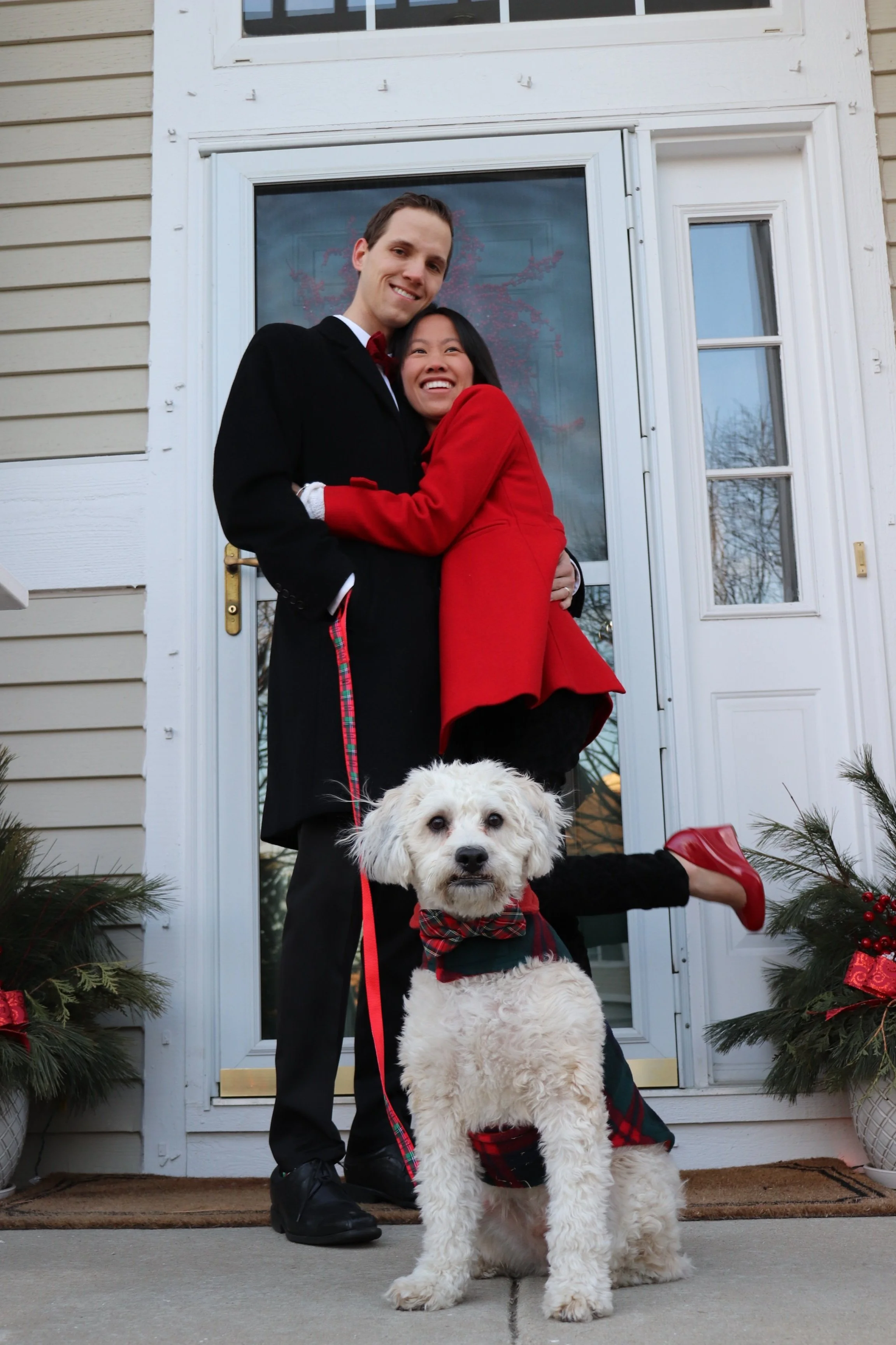 A smiling couple stands in front of a door, hugging each other, while a fluffy white dog in a plaid scarf sits in the foreground. The woman is wearing a red coat and red shoes, and the man is in a black coat.