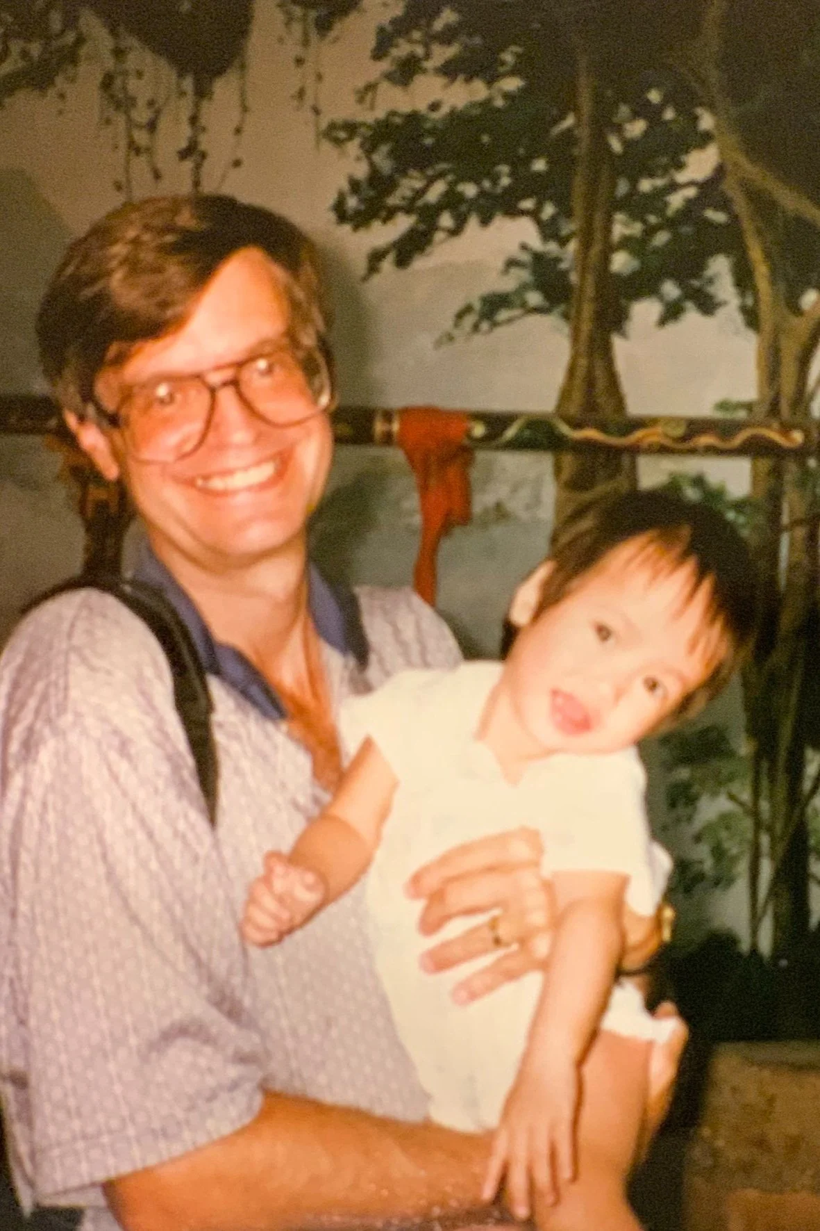 A man holding a baby with a tree-themed background.