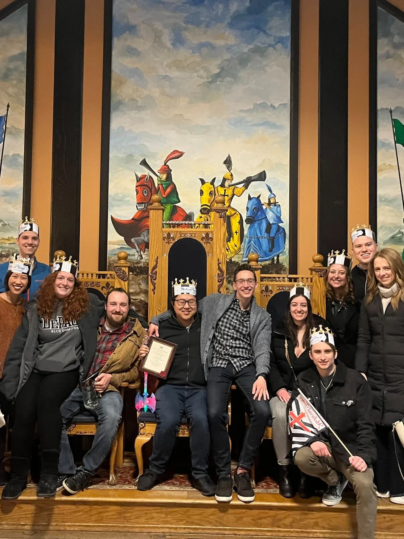 Group of people wearing medieval-style paper crowns, posing in front of a mural depicting knights on horseback, inside a themed restaurant with wooden thrones and a castle decor.