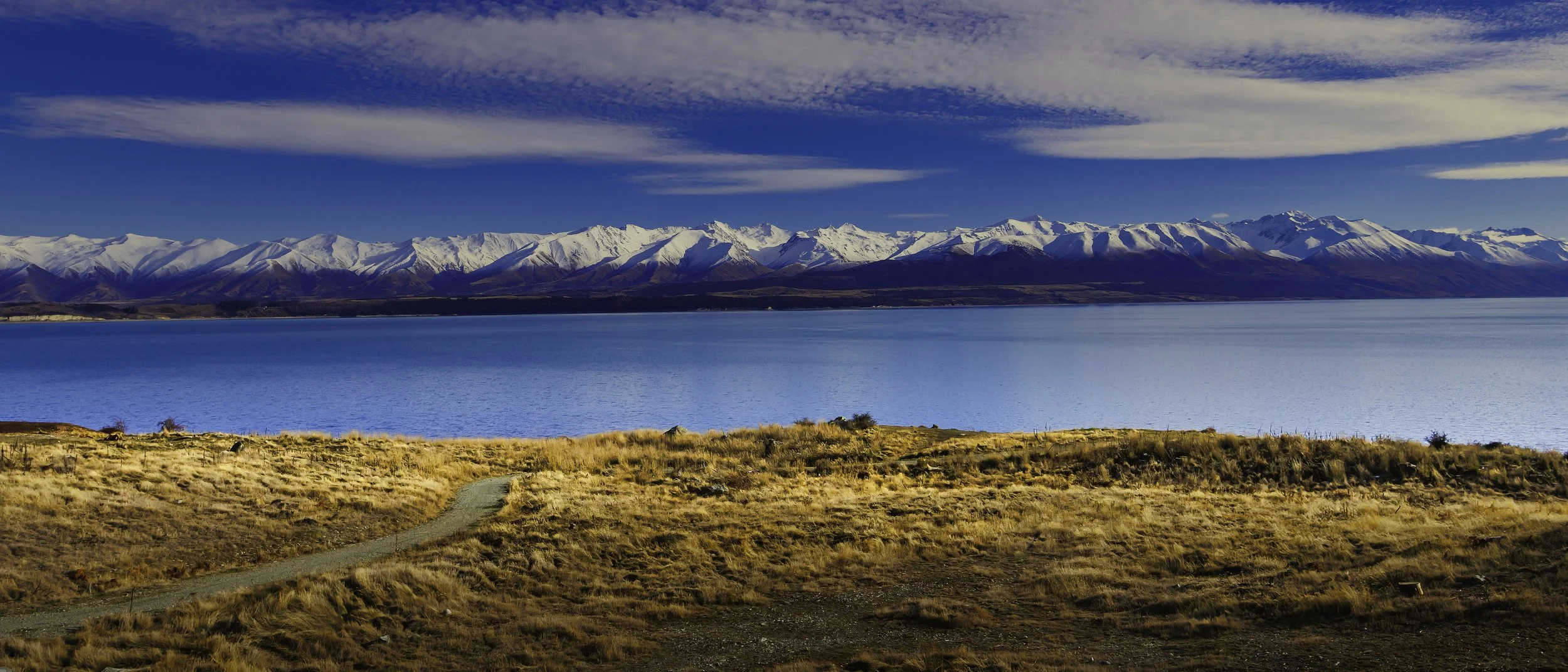 A landscape featuring a large, bright blue lake with snow-capped mountains in the background and a grassy foreground with a dirt path.