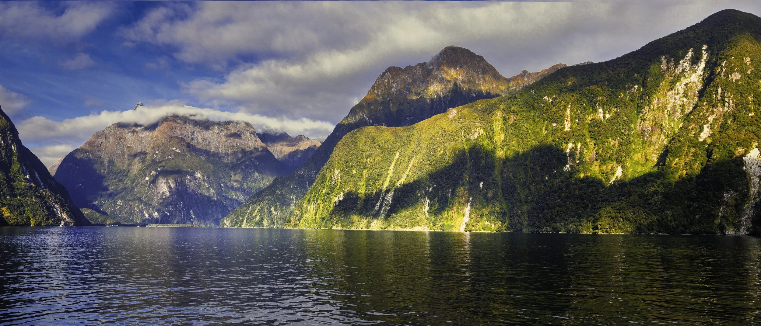 Scenic view of a large body of water with mountains covered in green foliage in the background. The mountains are partly in shadow, with some sunlight highlighting the slopes. The sky has clouds and patches of blue.