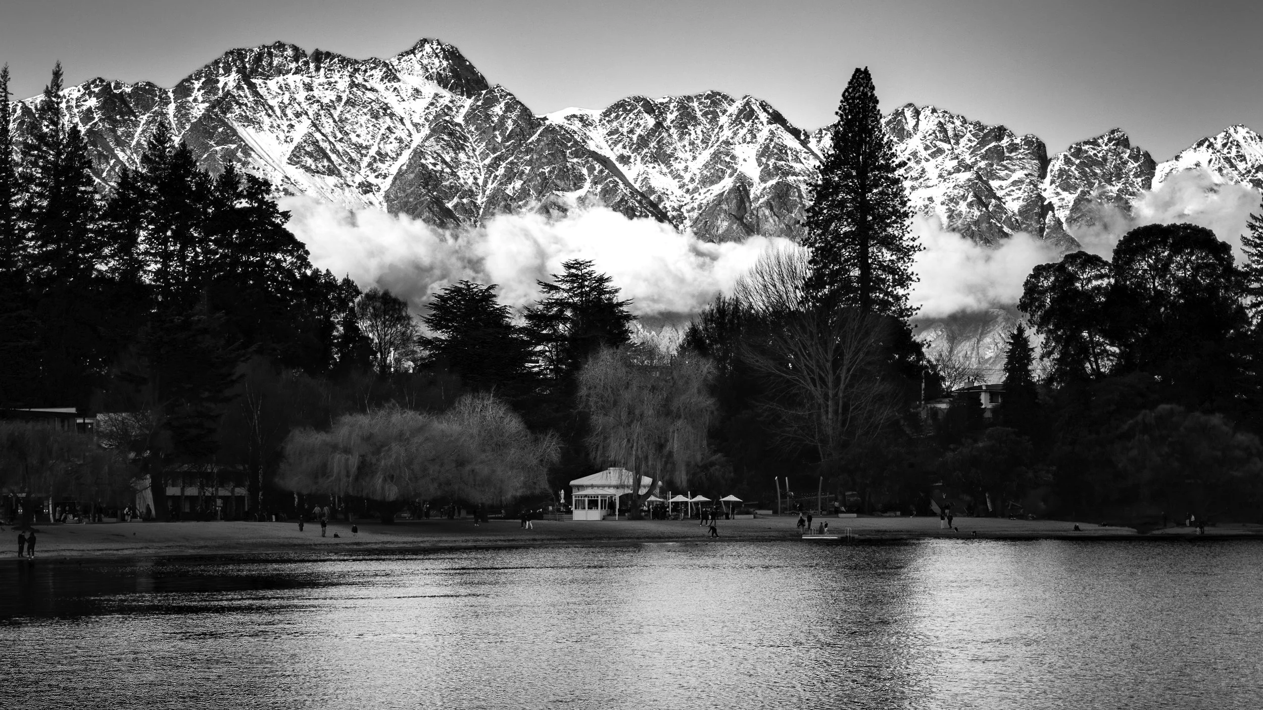 Black and white photo of Remarkable mountains with snow on their peaks, trees in the foreground, and a body of water at the bottom of the image.