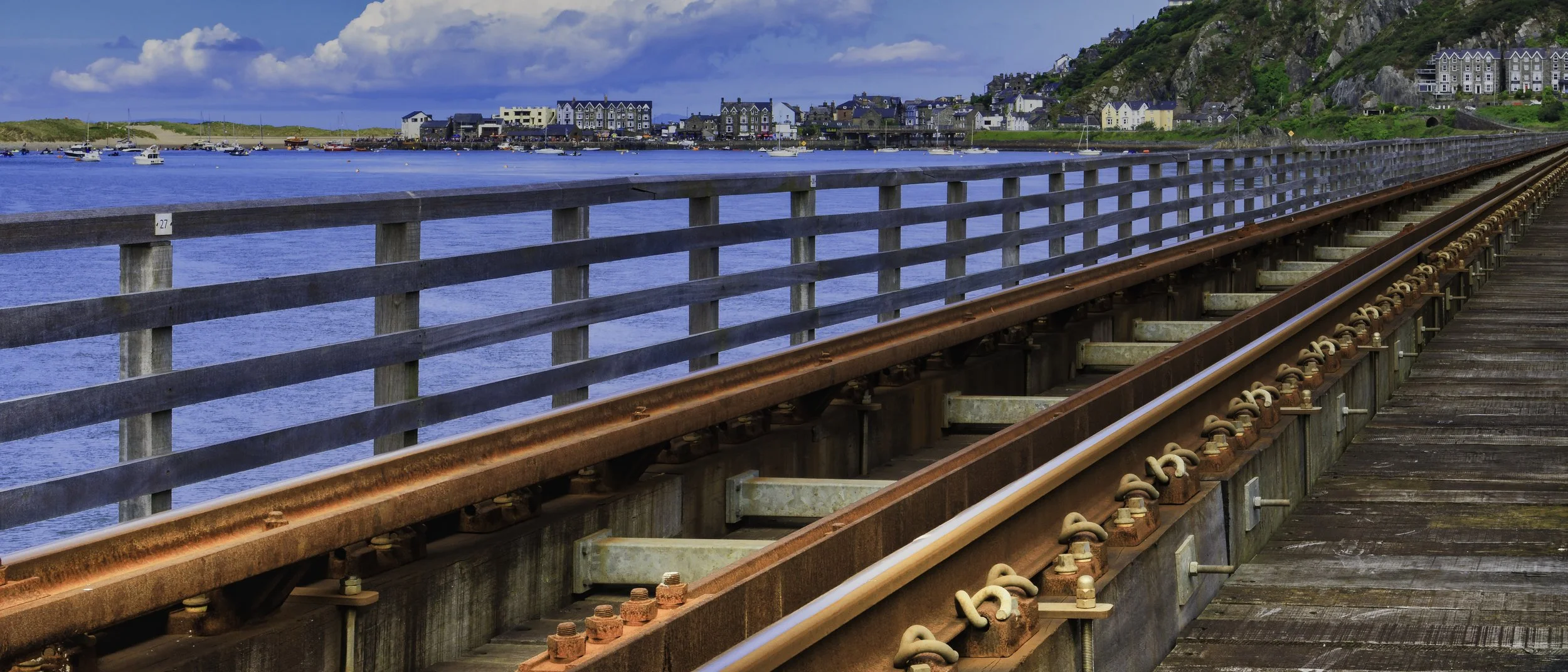 Railroad tracks on a wooden bridge near water, with a town and hillside in the background under cloudy sky.