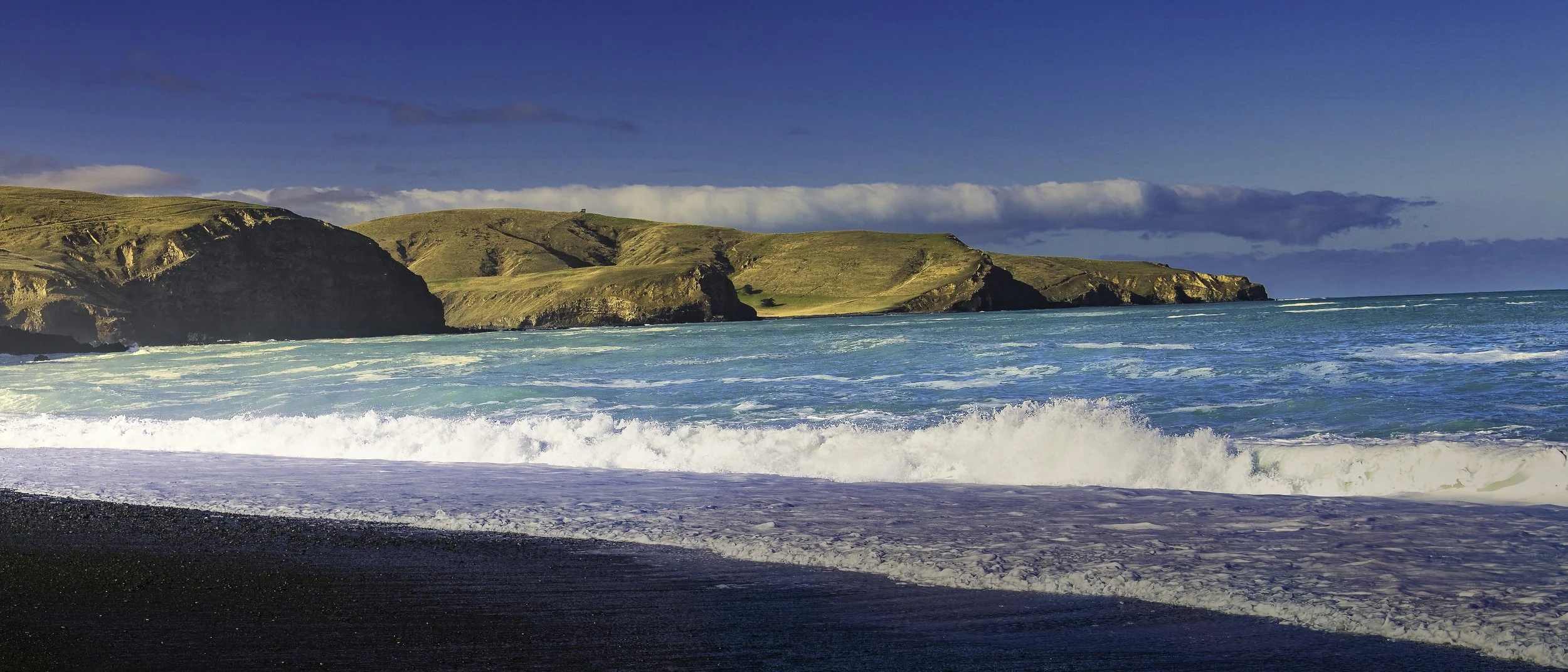 Coastal landscape with cliffs, grassy hills, and a blue ocean with waves crashing on a black sandy beach under a partly cloudy sky.