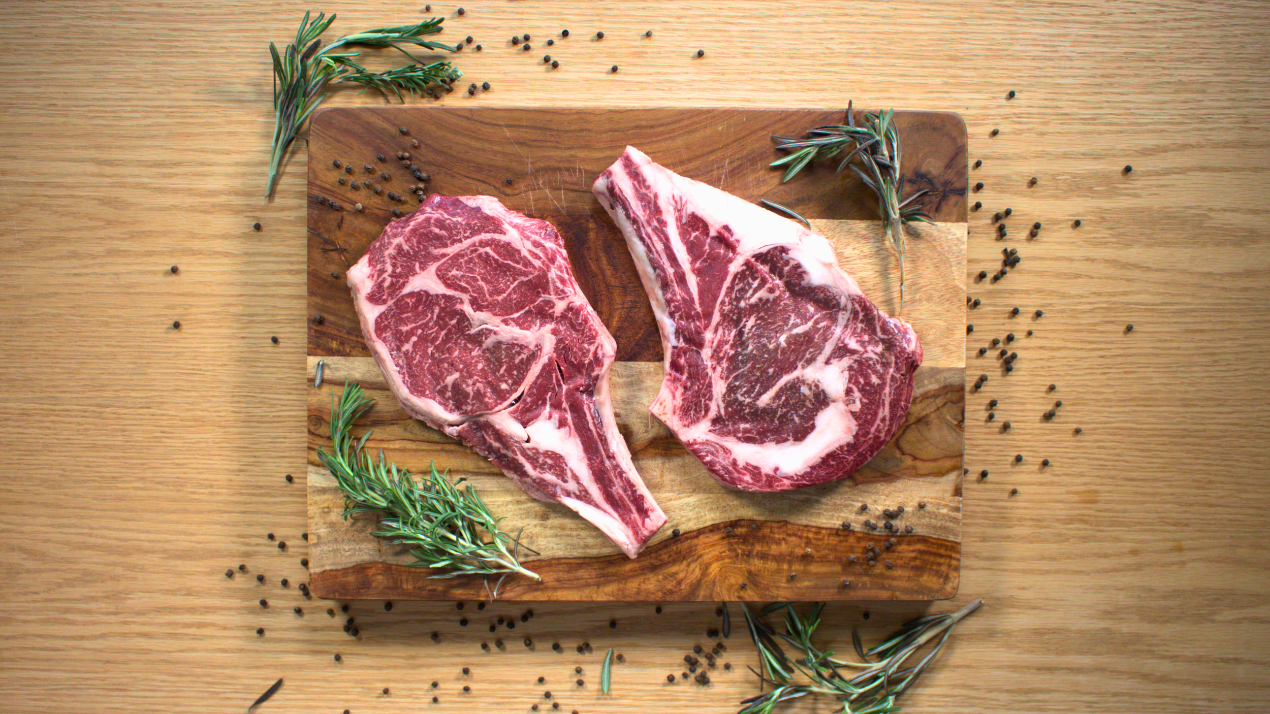 Two uncooked beef steaks with marbling on a wooden cutting board, surrounded by sprigs of rosemary and black peppercorns scattered on a wooden surface.
