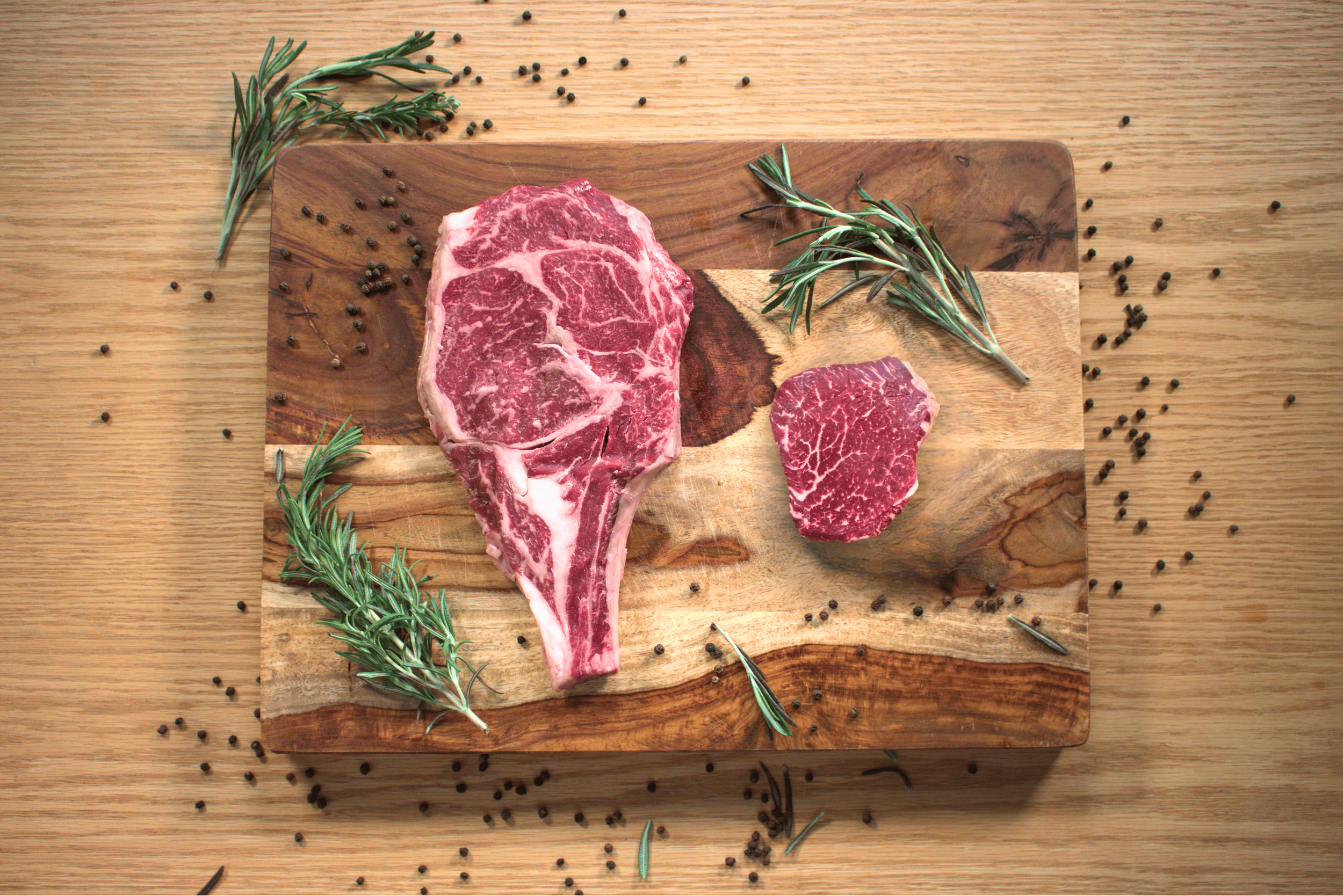 A raw T-bone steak and a slice of ribeye steak on a wooden cutting board, garnished with sprigs of rosemary and scattered black peppercorns.