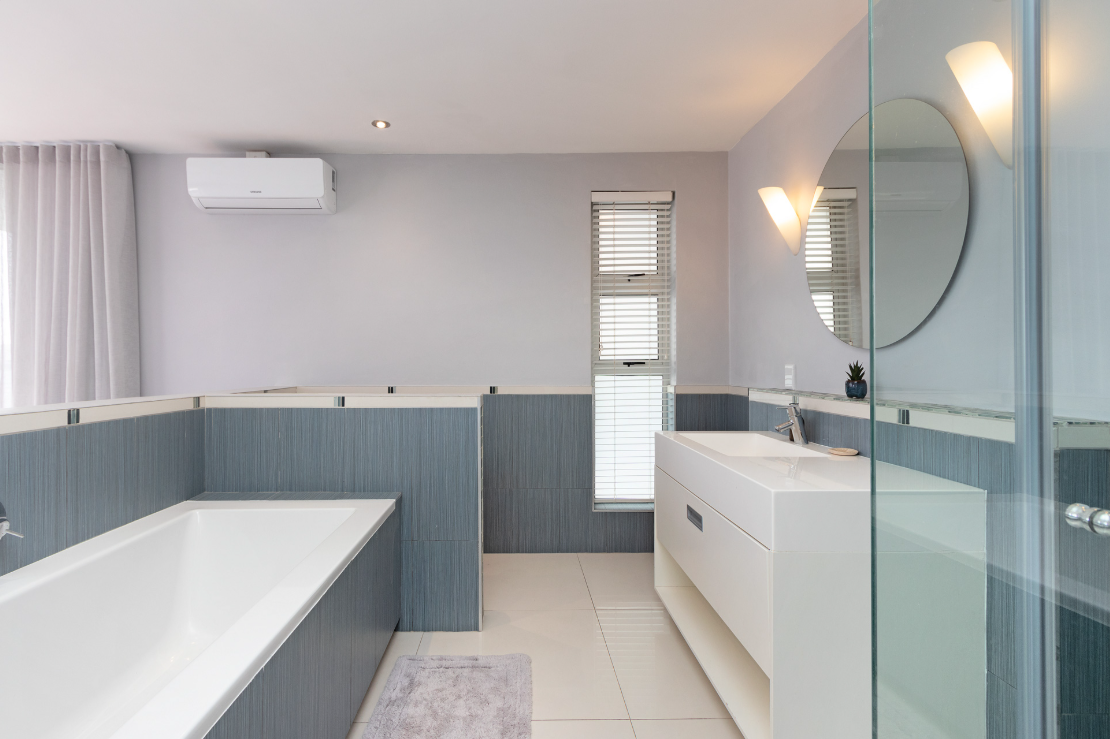 Modern bathroom with a white bathtub, a white vanity with a sink, a round mirror, a small potted plant, and windows with blinds.