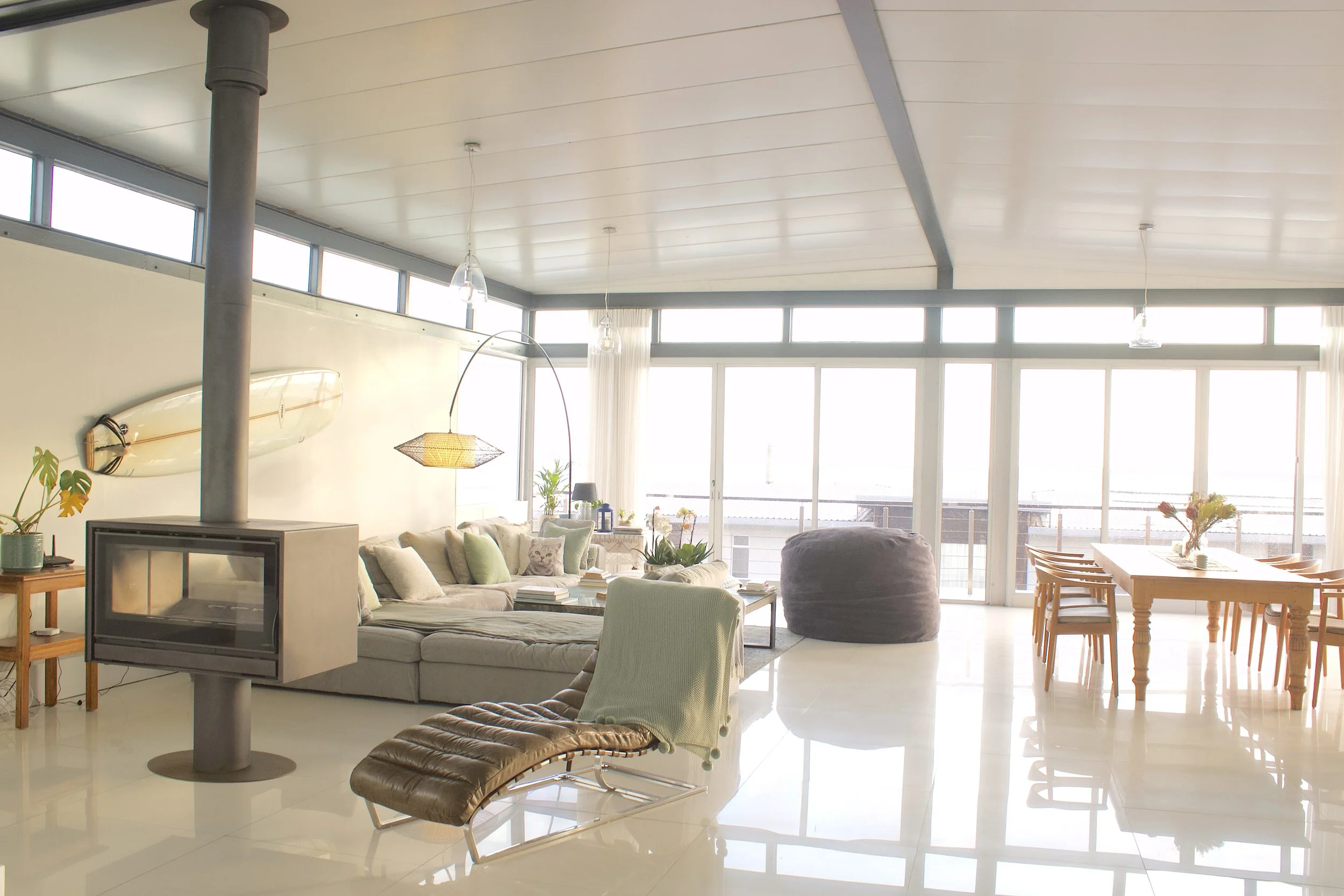 A spacious, modern living room with large windows letting in lots of natural light. The room has a light-colored ceiling and floors, with a mix of comfortable furniture including a sectional sofa, a lounge chair, and a wooden dining table with chairs