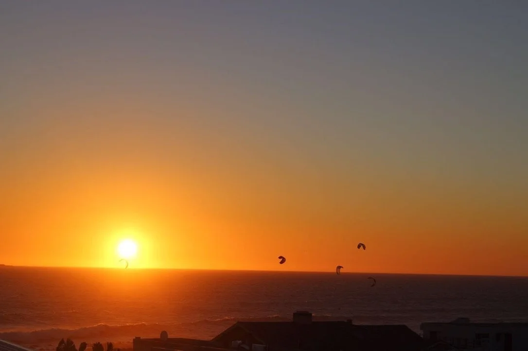 Sunset over the ocean with silhouetted houses in the foreground and four kite surfers visible in the water.