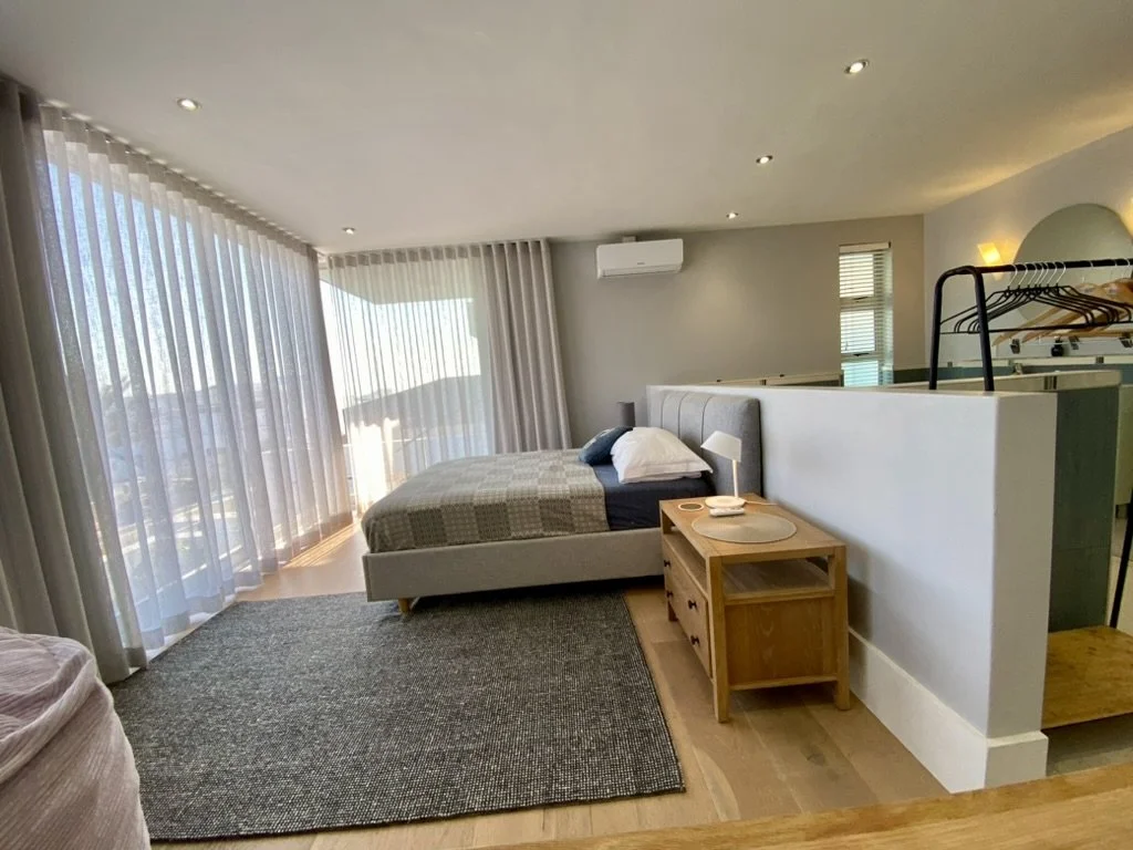 A modern queen-size bedroom with a large window covered by sheer curtains, a high density foam bed with pillows, a bedside table with a lamp and dishes, a gray carpet. There is an air conditioning unit on the wall and a small window with blinds.