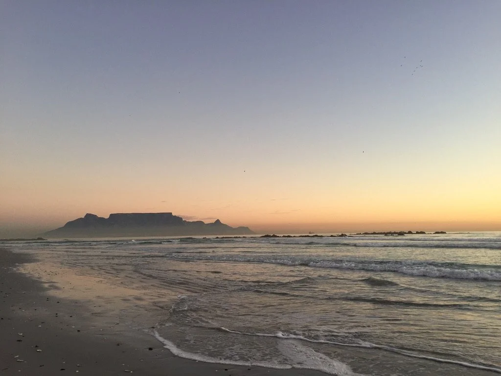 Sunset over a beach with calm waves and Table Mountain in the distance.