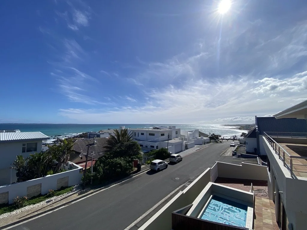 View of a coastal neighborhood of Bloubergstrand with houses, parked cars, a street, and the ocean in the background under a bright blue sky with some clouds and the sun shining.
