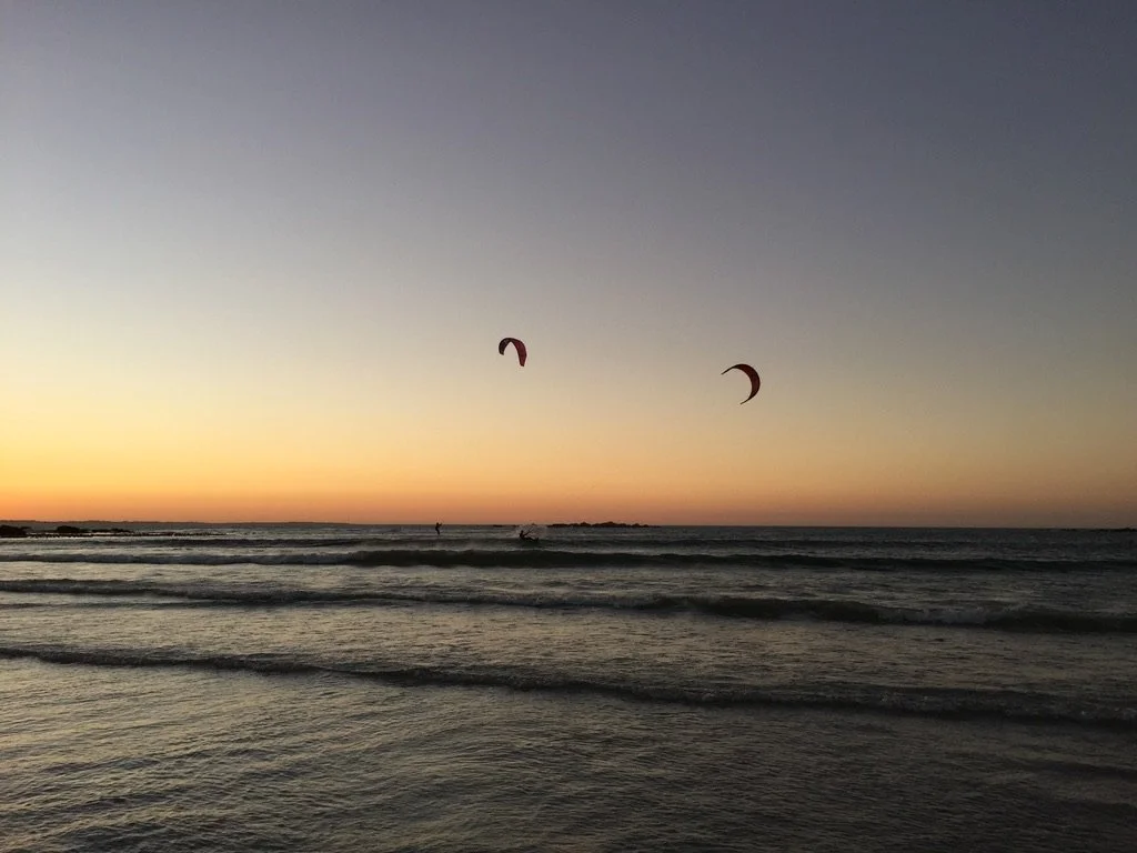 Sunset over the ocean with two kitesurfers riding the waves in the distance.