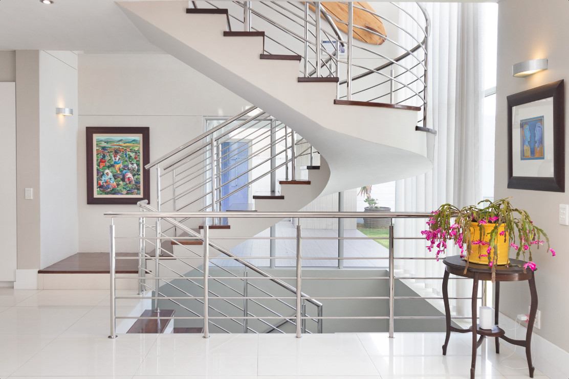 Interior view of a modern house with a curved staircase featuring metal railings, white walls, wall-mounted lights, colorful artwork on the walls, a wooden table with a yellow pot of pink flowers, and large windows with vertical blinds.