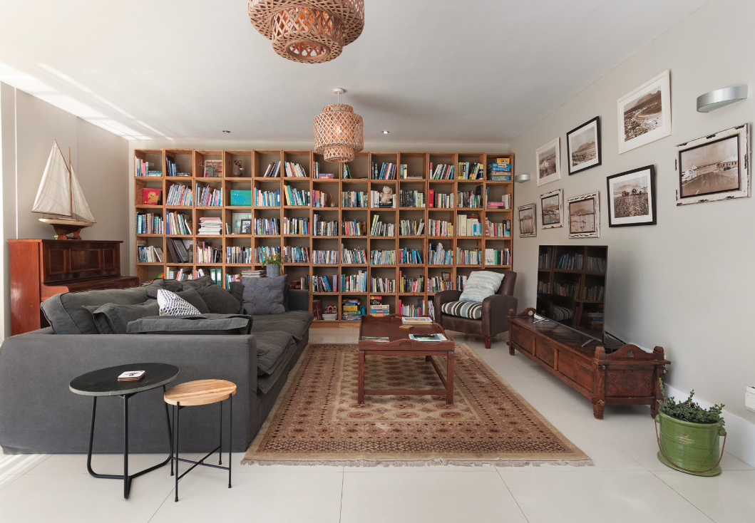 Living room with a large wooden bookshelf filled with books, a dark gray sofa, striped armchair, wooden coffee table, flat-screen TV on a wooden stand, vintage picture frames on the wall, a potted plant, and nautical decor including a model sailboat.