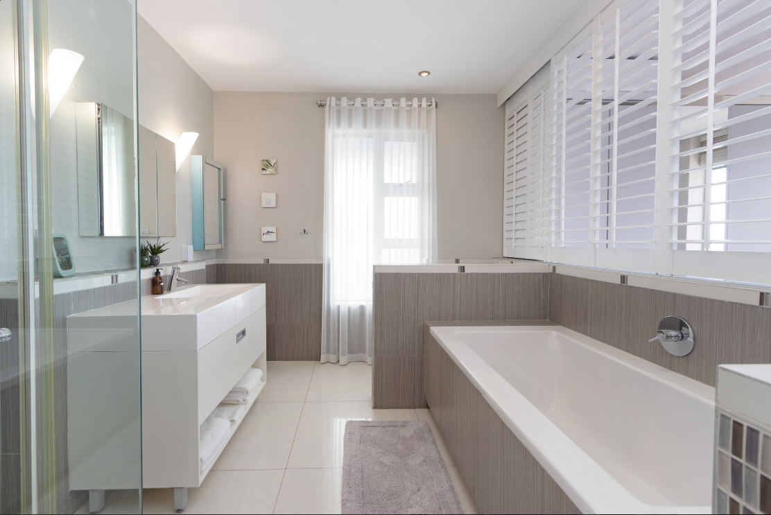 A modern bathroom with a white bathtub, a sink vanity, large windows with white shutters, and light-colored tiles.