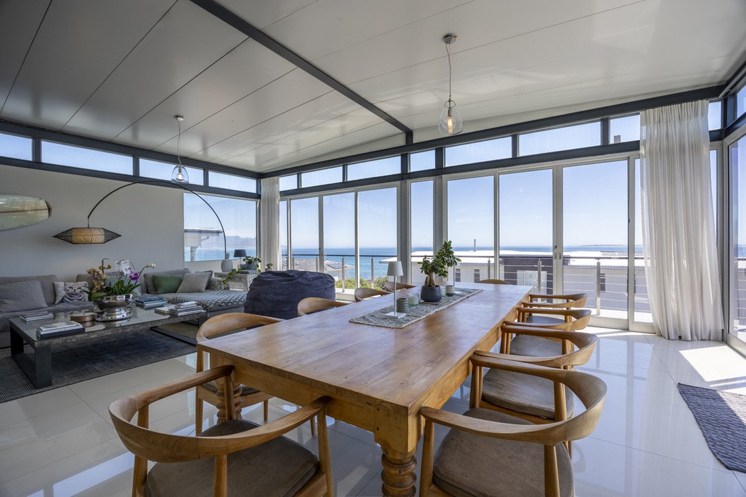 Sunlit dining area with a wooden table and six chairs, overlooking an ocean view through large glass windows.