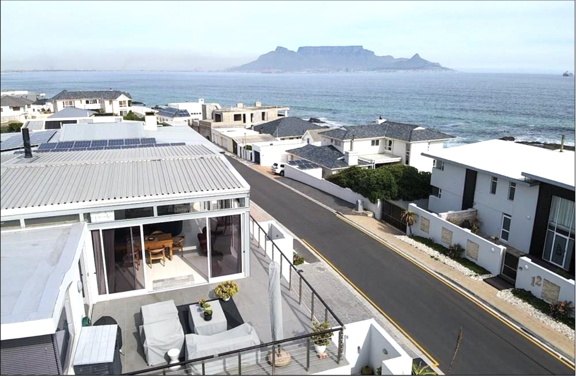 View of residential houses by the ocean with Table Mountain in the background, some rooftops have solar panels, and the scene is viewed from a balcony.