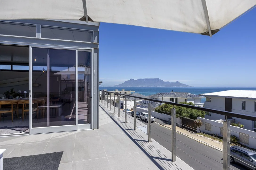 Balcony with glass door and railing overlooking a coastal town and Table Mountain under a clear blue sky.