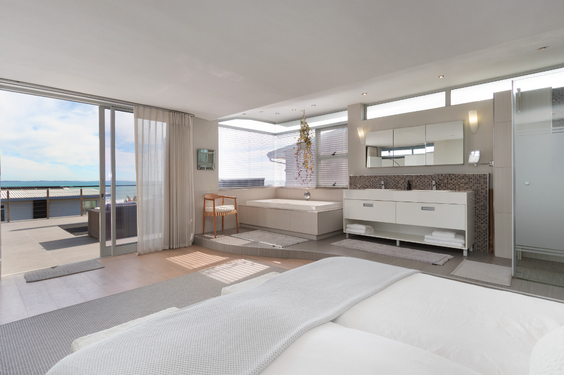 Modern king bedroom with a large sliding glass door leading to an outdoor patio with ocean view, white walls and ceiling, a king bed with white linens, an ensuite bathroom area with a vanity, mirror, and bathtub, and sheer curtains.