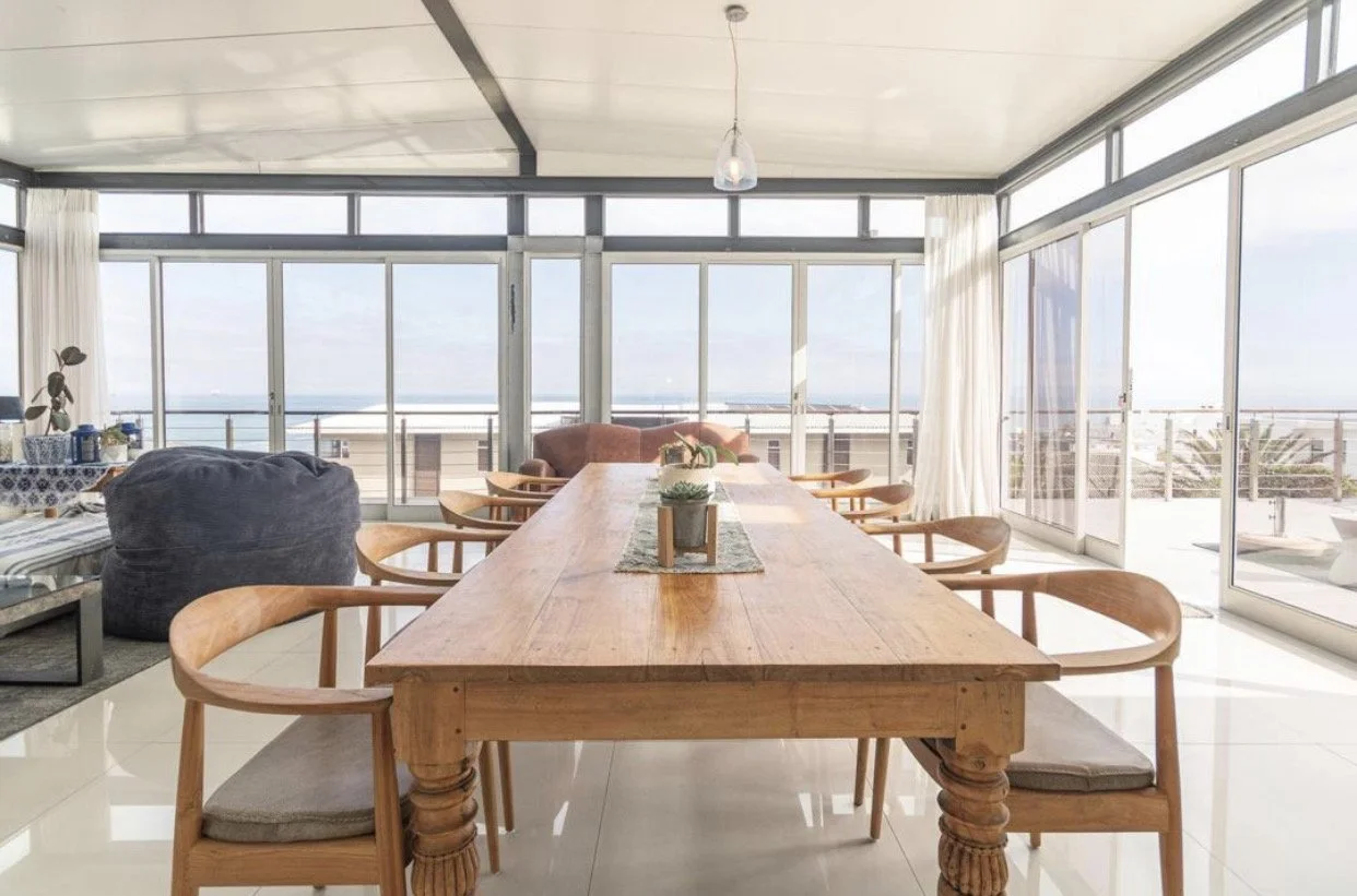 Bright dining room with a large wooden table, 10 chairs, and floor-to-ceiling windows overlooking Robben Island, Table Mountain and the ocean.