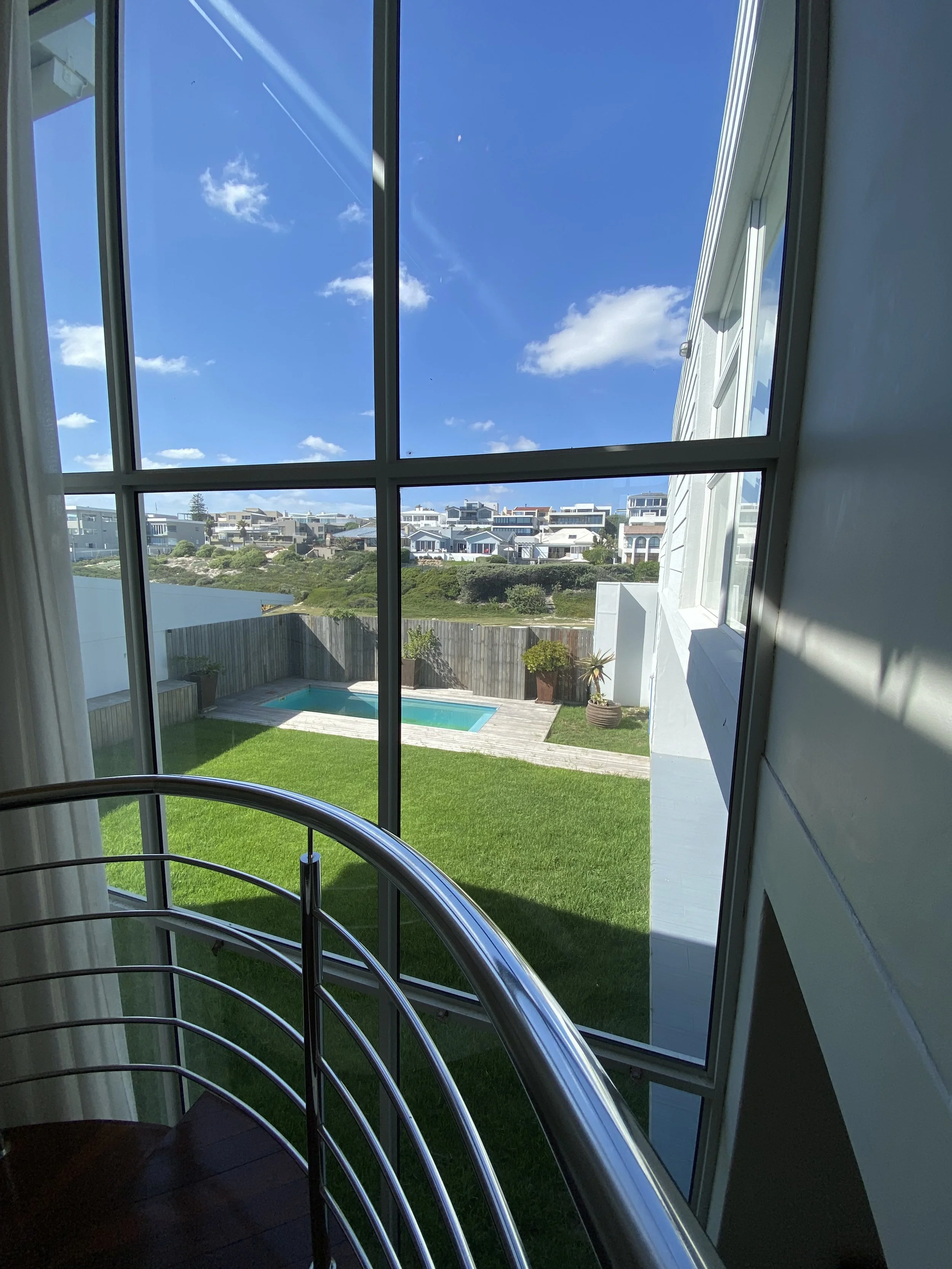 Looking out a large window at a backyard with a swimming pool, surrounded by a grassy lawn and wooden fencing, with modern houses and a blue sky with some clouds outside.