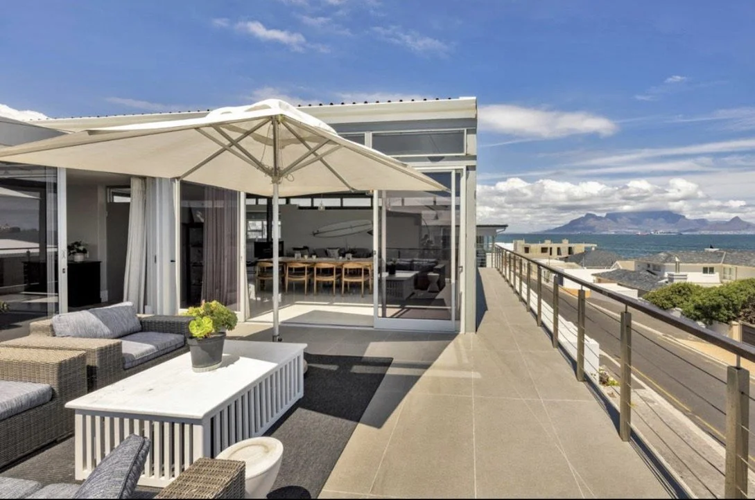 Balcony view of outdoor seating with sofa, table, and umbrella overlooking beach and ocean under blue sky.