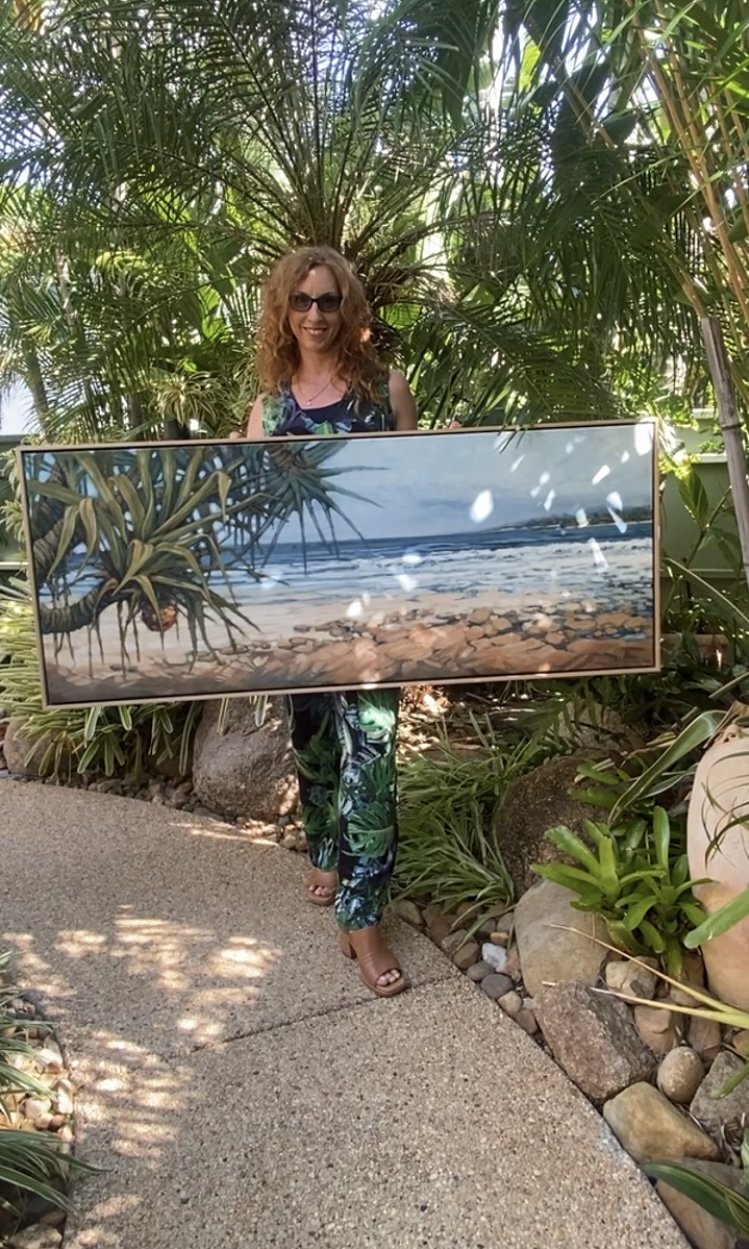 A woman with curly hair and sunglasses standing outdoors on a garden path, holding a large framed painting of a beach scene with rocks, ocean, and sky, surrounded by lush green tropical plants.