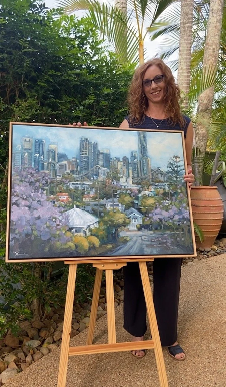 Woman with curly red hair and glasses standing outdoors holding a painting of a cityscape with tall buildings and flowering trees.