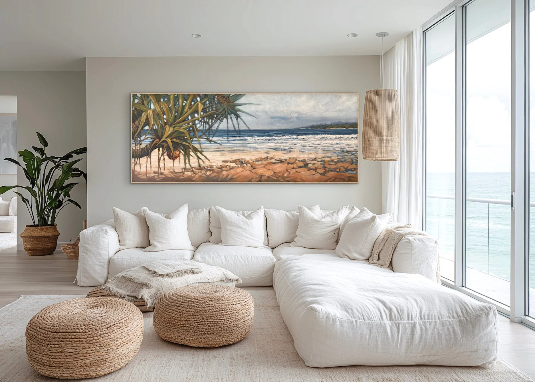 Living room with a white sofa, large wall art of a beach scene, potted plant, woven poufs, and floor-to-ceiling windows showing the ocean.