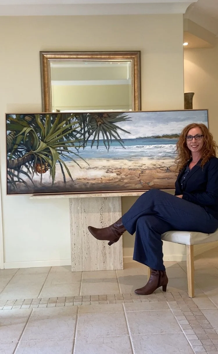 A woman with curly red hair and glasses sitting on a chair in front of a landscape painting of a beach with ocean waves, sandy shore, and cloudy sky, in a room with tiled floor and a mirror on the wall.