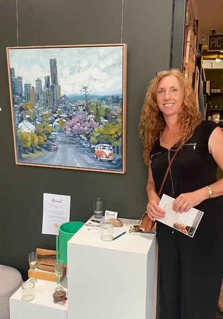 The Artist with curly red hair smiling, holding a brochure, standing next to the painting that was a finalist in an art competition. Behind her is a framed painting of a Brisbane cityscape with Jacarandas, cars, and the Story Bridge.