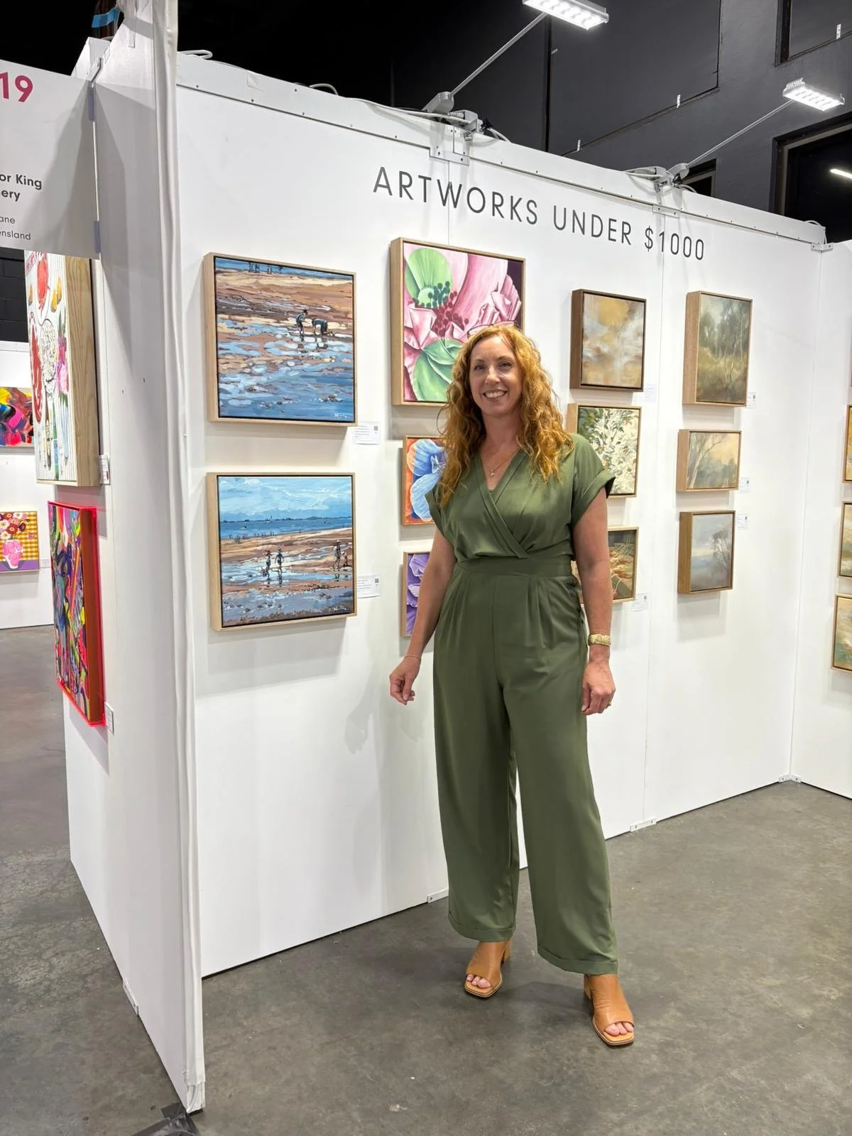 The female Artist with curly red hair and a green jumpsuit, standing in front of an art display at the Brisbane Affordable Art Fair smiling at the camera.