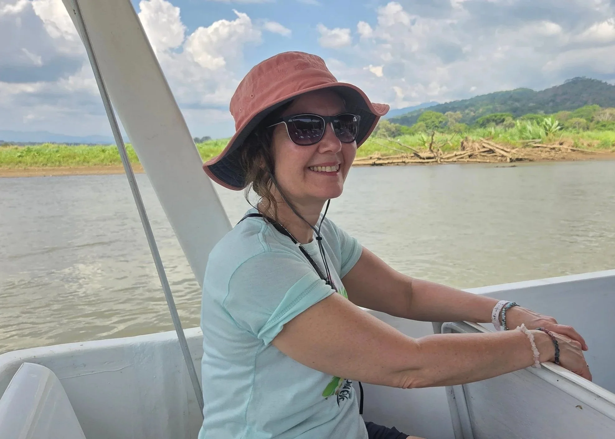 Nichola smiling while sitting on a boat, wearing a pink wide-brimmed hat, sunglasses, and a light blue shirt, with a river, trees, and hills in the background.