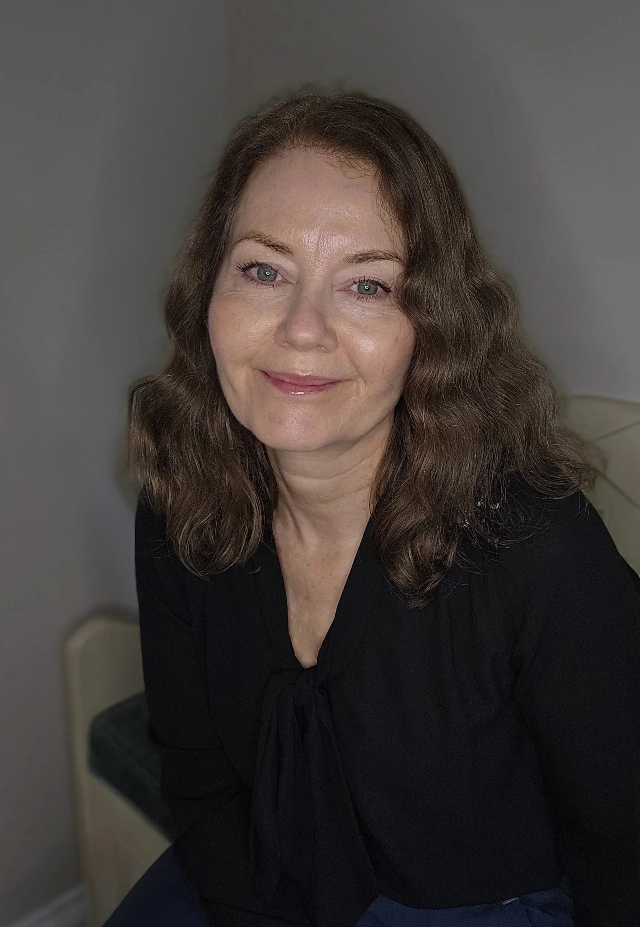 A woman with wavy brown hair and blue eyes, smiling, wearing a black top, sitting indoors with a blurred neutral background.