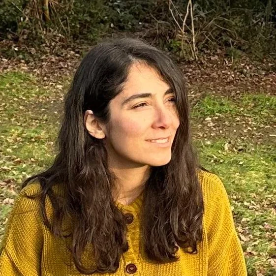 Junge Frau mit braunen, lockigen Haaren in einer gelben Jacke sitzt im Freien auf einer Wiese mit Bäumen im Hintergrund.