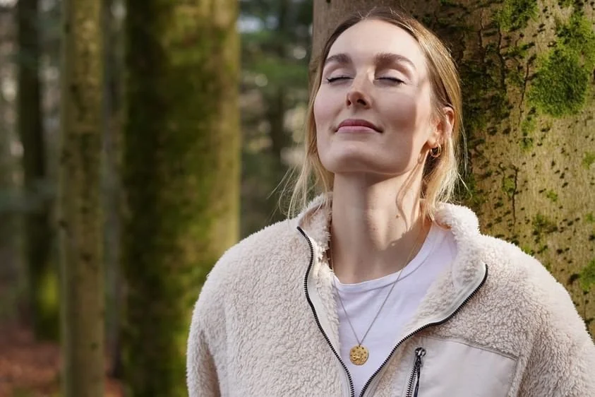 Eine junge Frau mit blonden Haaren, die die Augen geschlossen hat und entspannt in der Natur steht, neben einem Baum.
