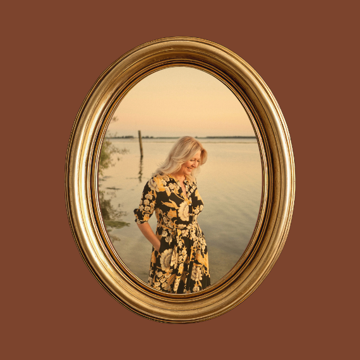 Een vrouw met blond haar in een bloemenjurk staat voor een spiegel met een uitzicht op een waterlandschap bij zonsondergang.