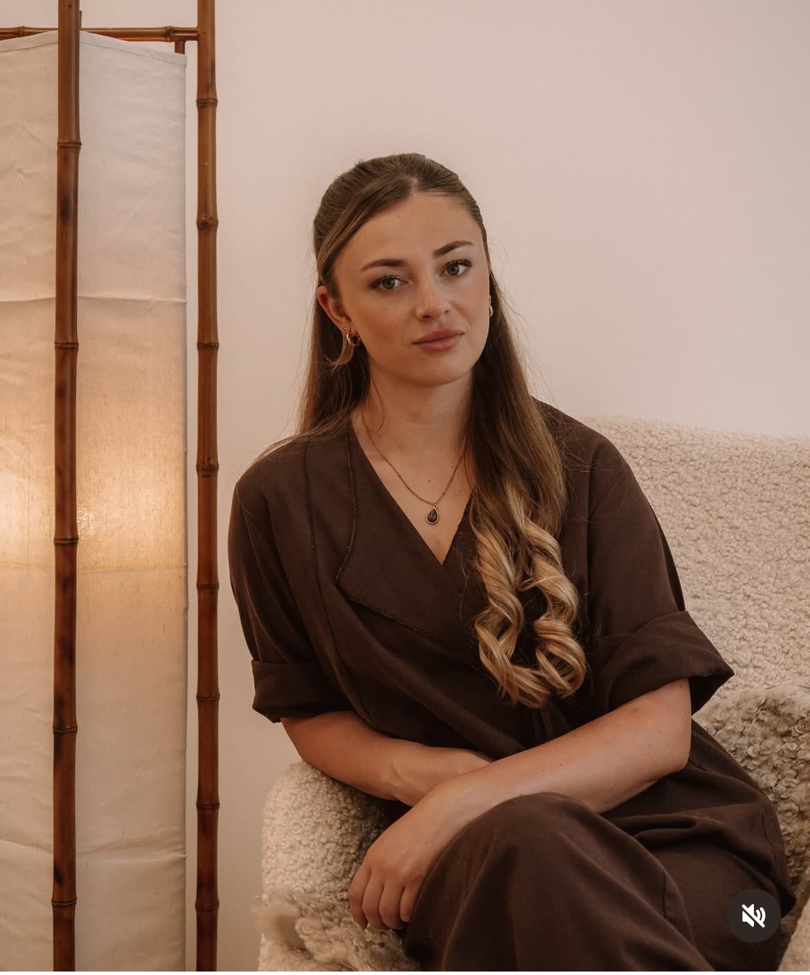 Een jonge vrouw met lang, golvend bruin haar, zittend op een beige, zachte stoel, gekleed in een bruin outfit, met een neutrale gezichtsuitdrukking, in een rustgevende binnenruimte met een houten kamerscherm op de achtergrond.