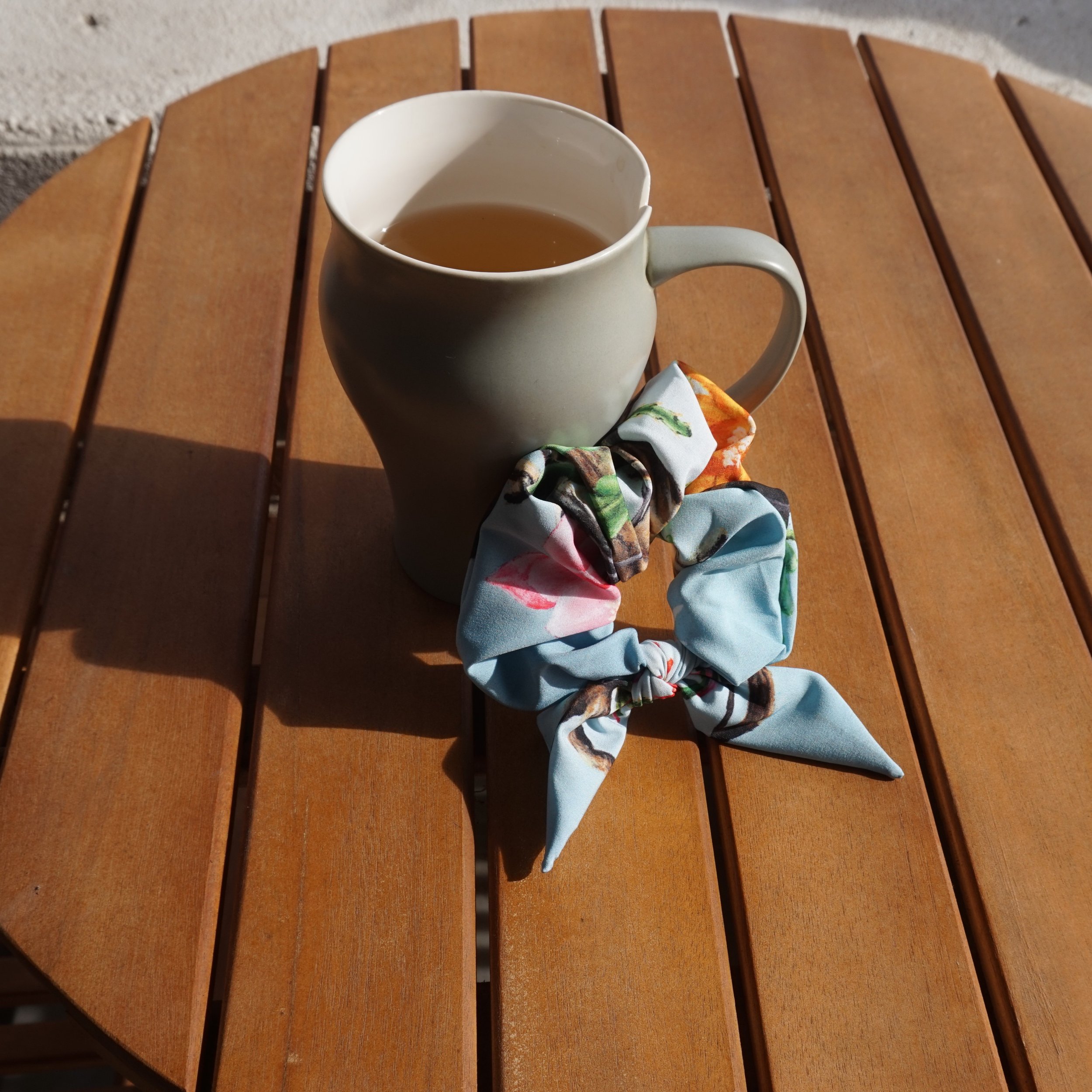 A young woman with brown hair and bangs, wearing a blue patterned scarf, white jacket, and black top, standing outdoors in front of a house with a porch and garden.