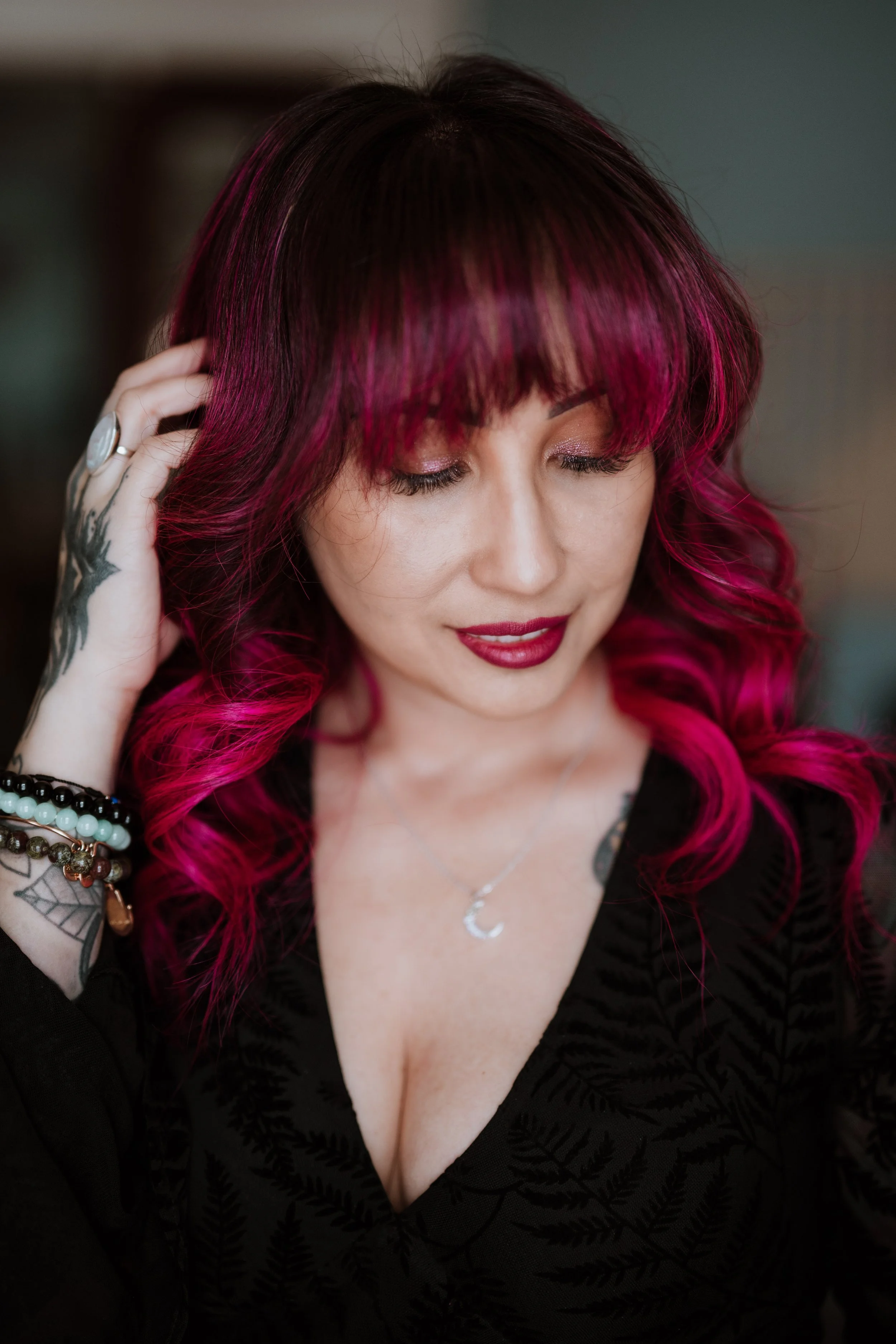 A woman with pink and black hair, wearing makeup with red lipstick, a black patterned top, and jewelry, touching her hair with her eyes closed.