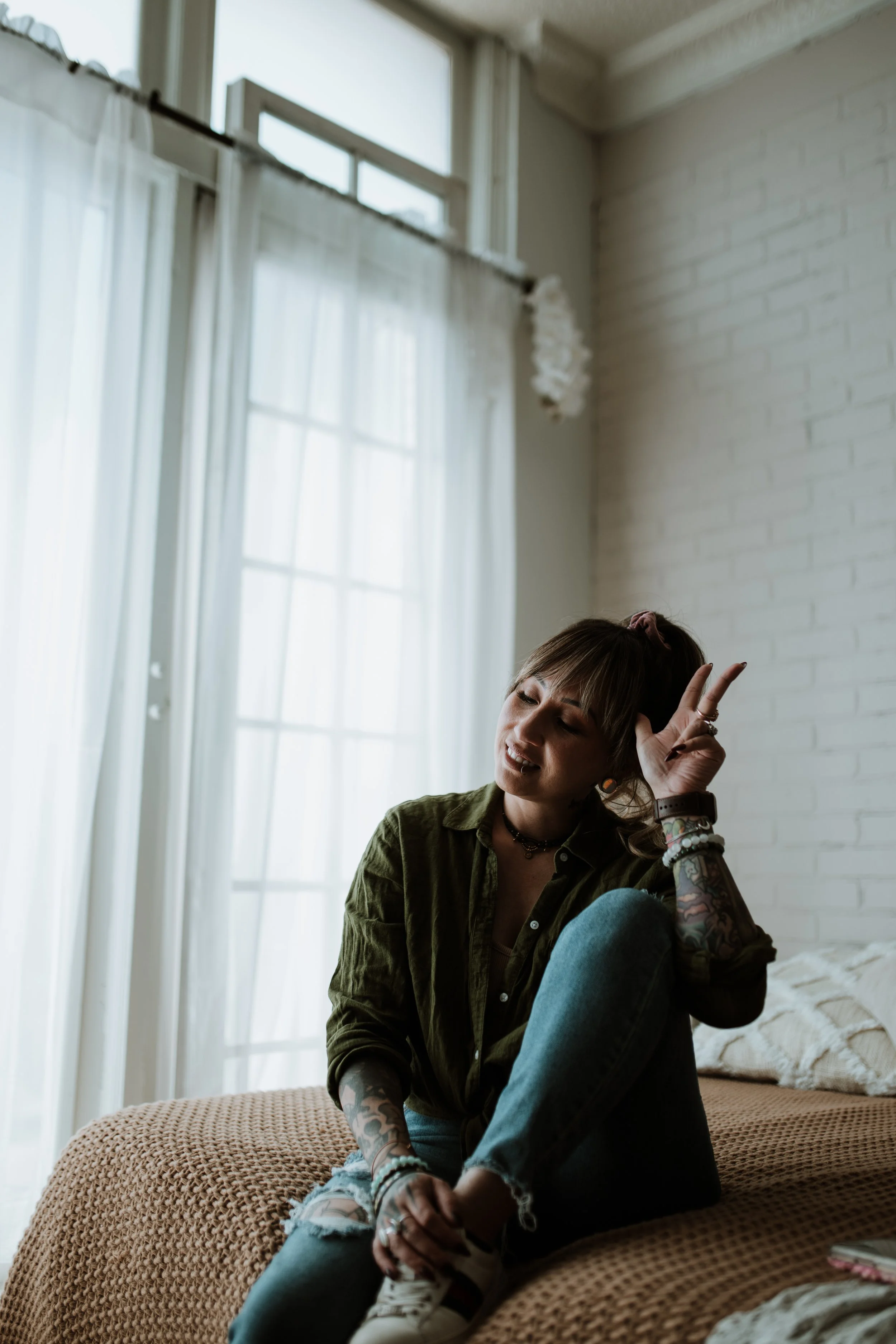 A woman with tattoos, wearing a green shirt and ripped jeans, sitting on a bed in a well-lit room with white curtains and a white brick wall, making a peace sign with her right hand.