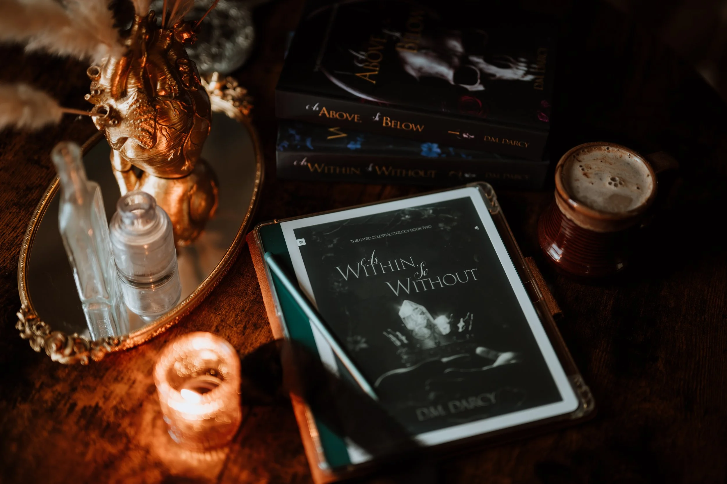 Kindle on table. Book titled As Within, So Without on screen. Stack of books beside it, titled As Above, So Below and As Within, So Without. Gold decor surrounds the works including a burning tealight candle, antique glass jars, and a mug of chai.