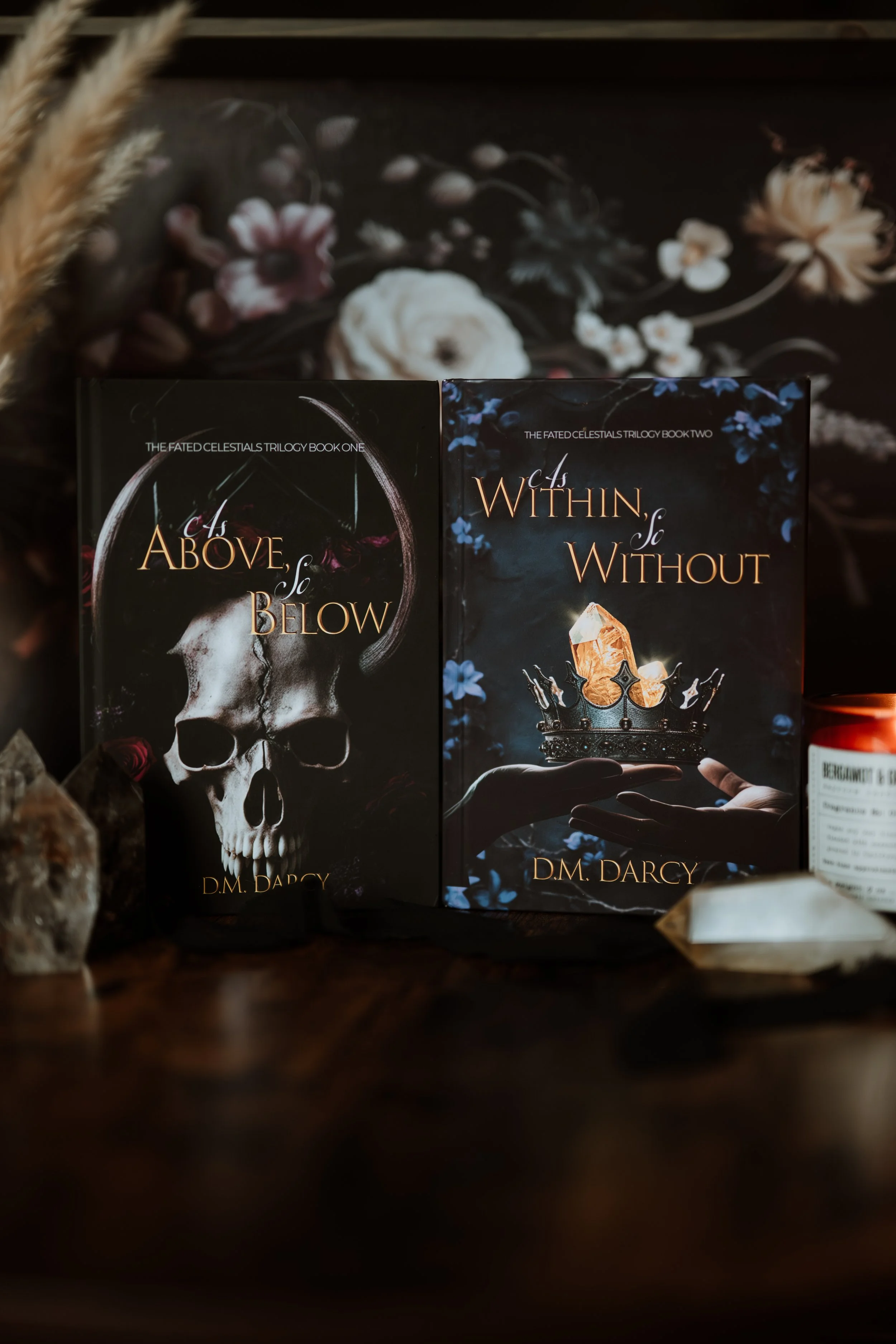Two books from The Fated Celestials Trilogy by D.M. Darcy, titled "As Above, So Below" with a skull on the cover and "As Within, So Without" with a crown and crystals on the cover, are displayed on a dark surface with crystals and a floral background.