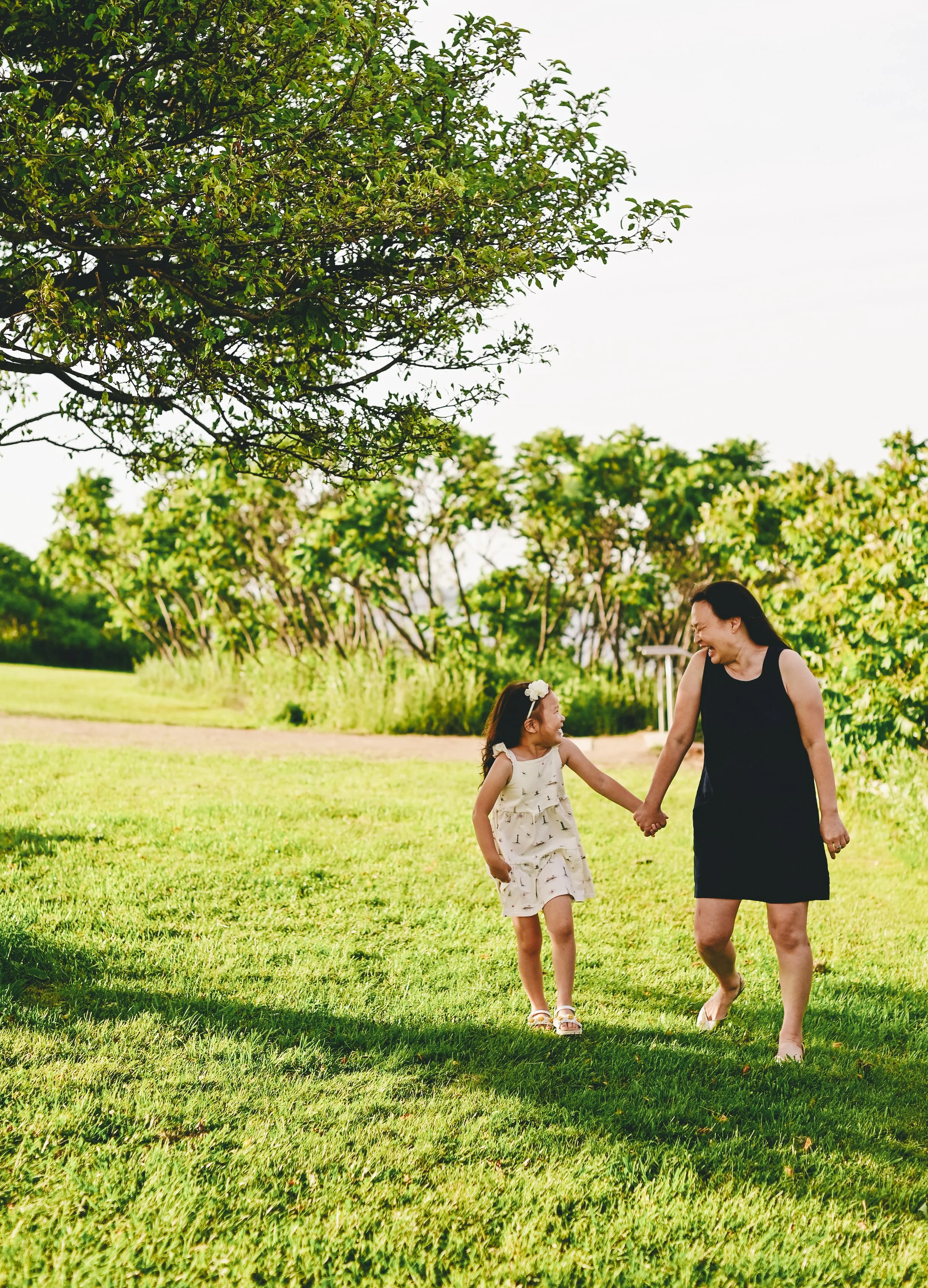 A mother and daughter holding hands and smiling while walking on grass on a sunny day.