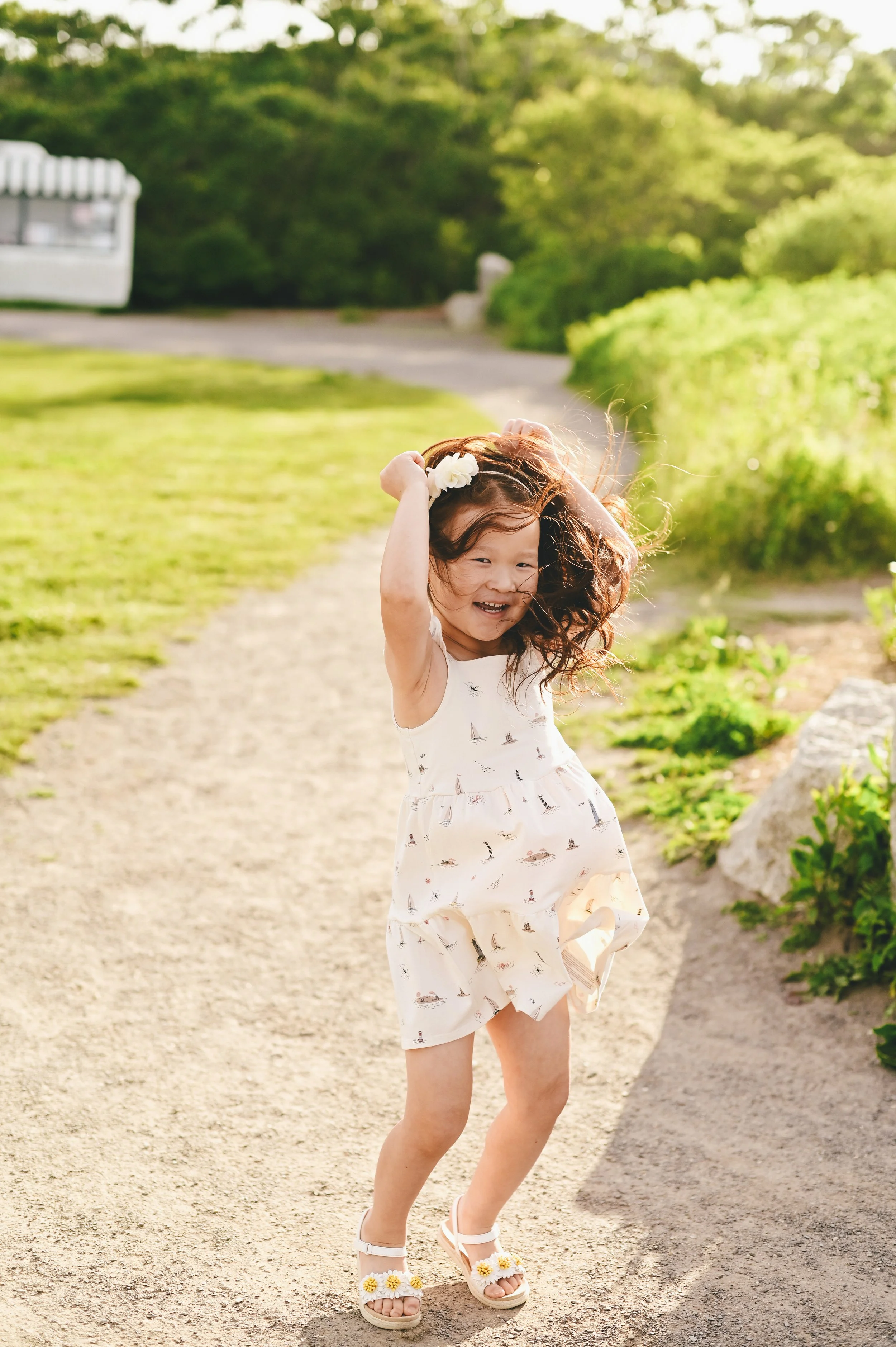 A young girl with wavy hair mid-jump, smiling and playing outdoors on a sunny day, wearing a white dress with sailboat prints.