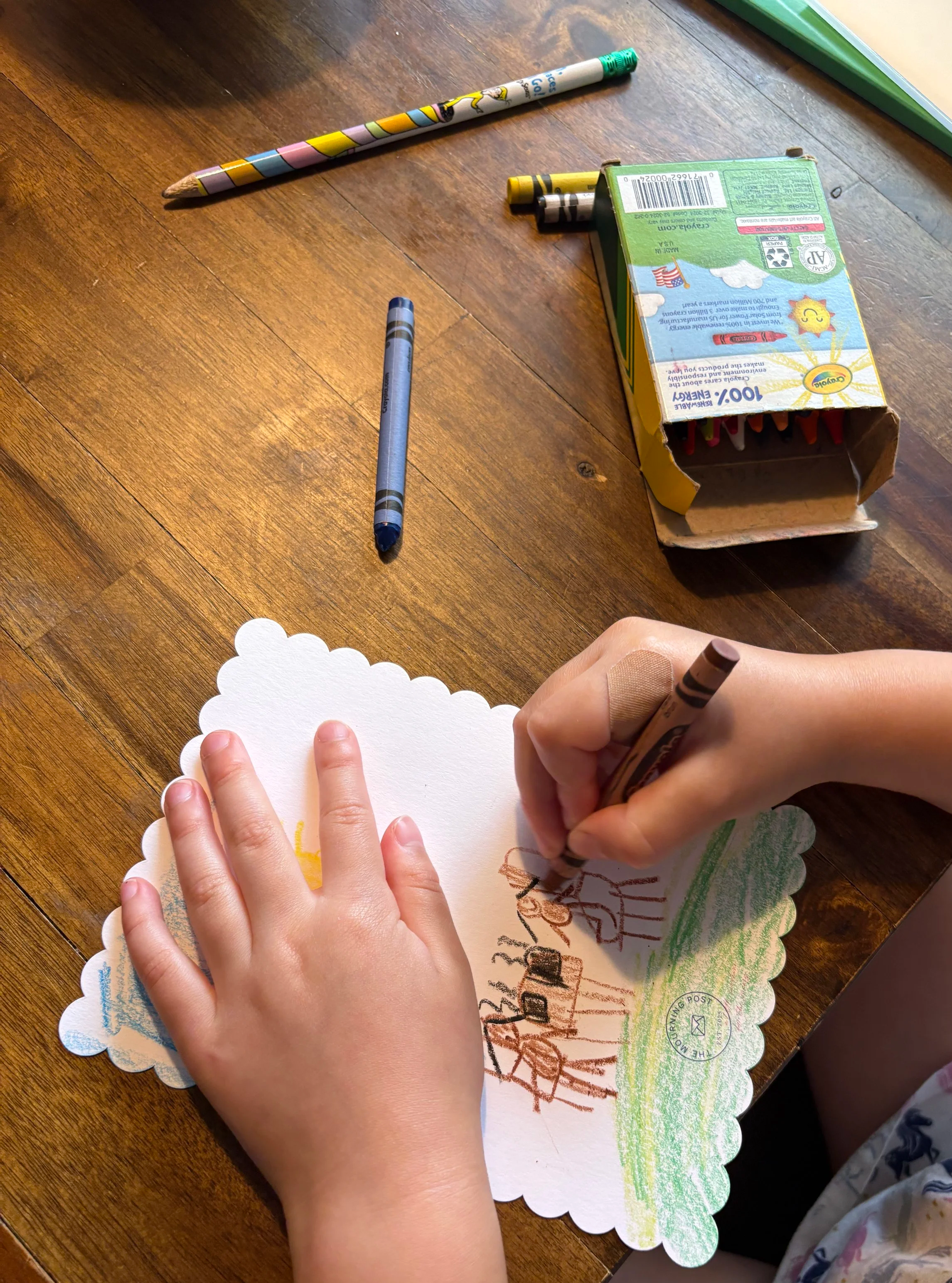 A child sits at a table drawing a colorful picture with crayons on a small note card